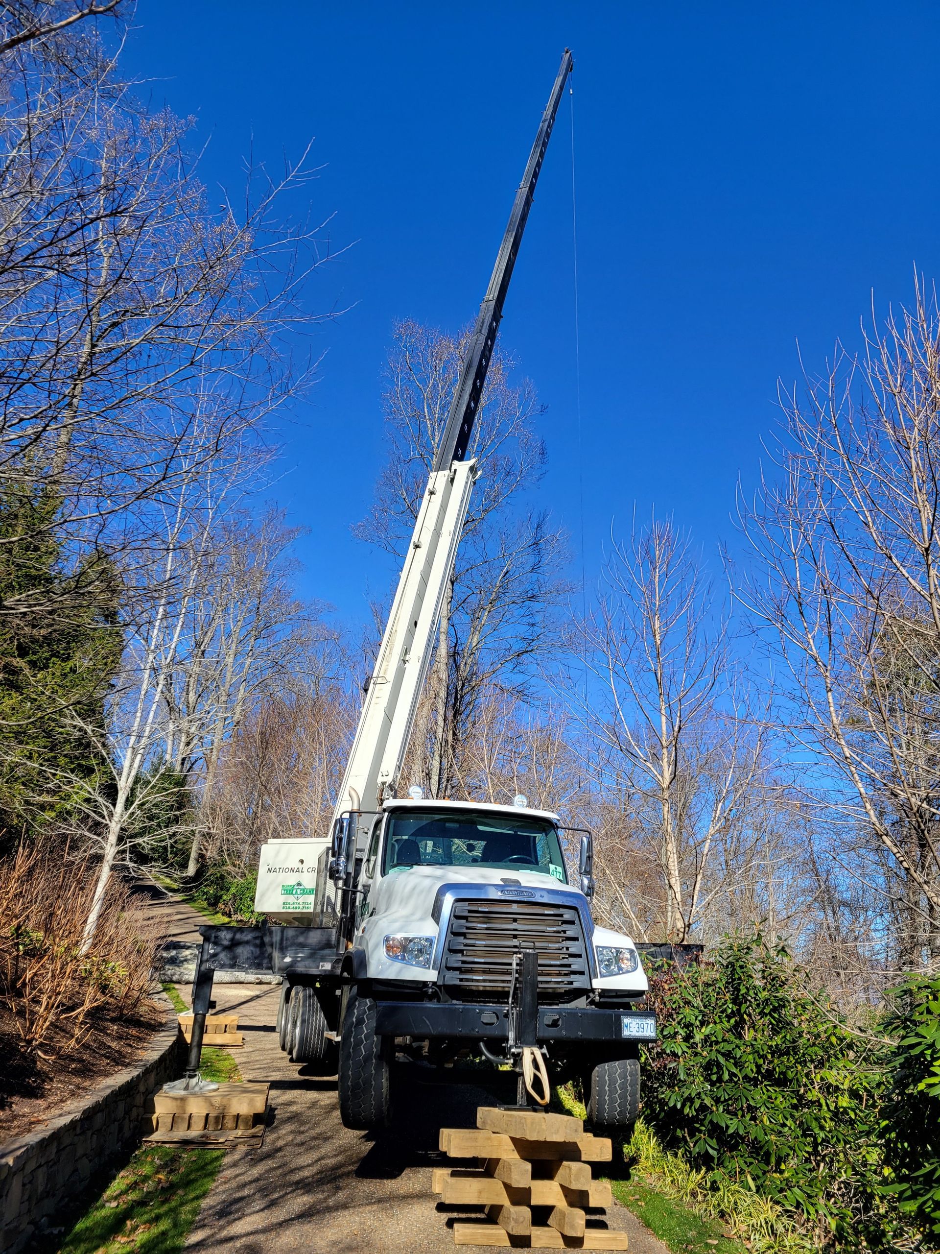 Large white truck with extended crane under a blue sky, on a wooded path.