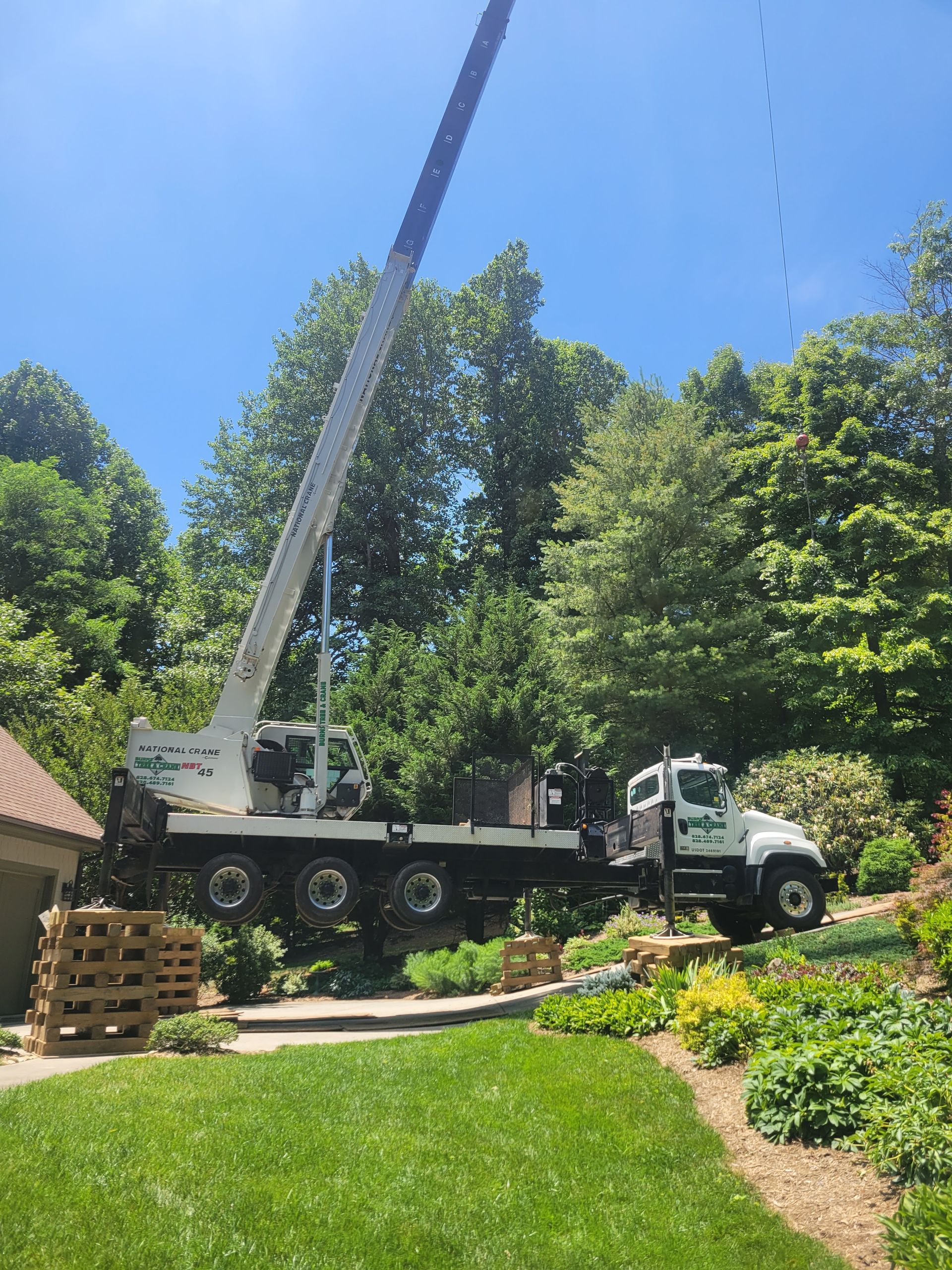 Crane truck lifting something, likely tree trimming, near a house with lush landscaping under a blue sky.
