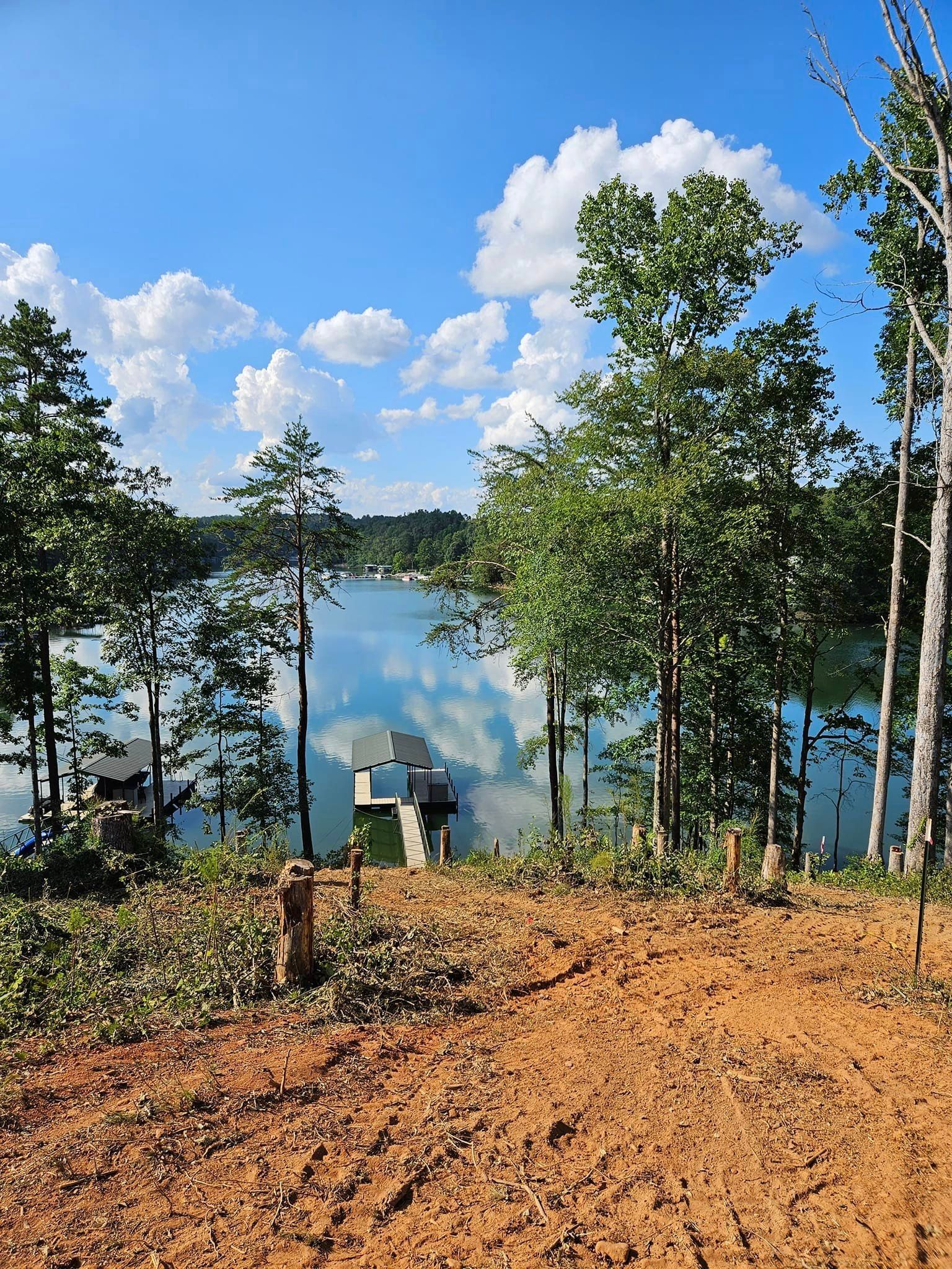 Lakeside view with blue sky and fluffy clouds. Trees frame the water, with a dock visible in the distance.