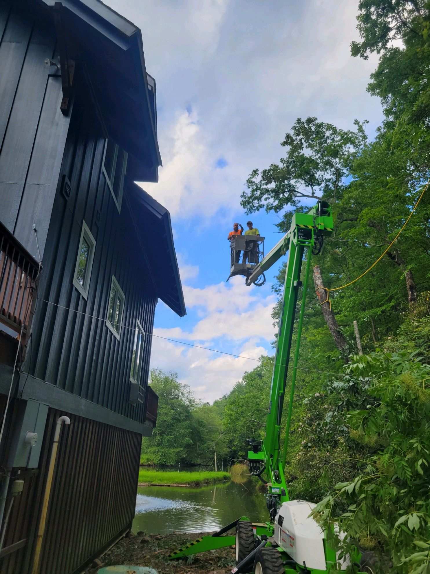 Workers in a green lift trim a tree next to a dark-sided building and pond on a cloudy day.