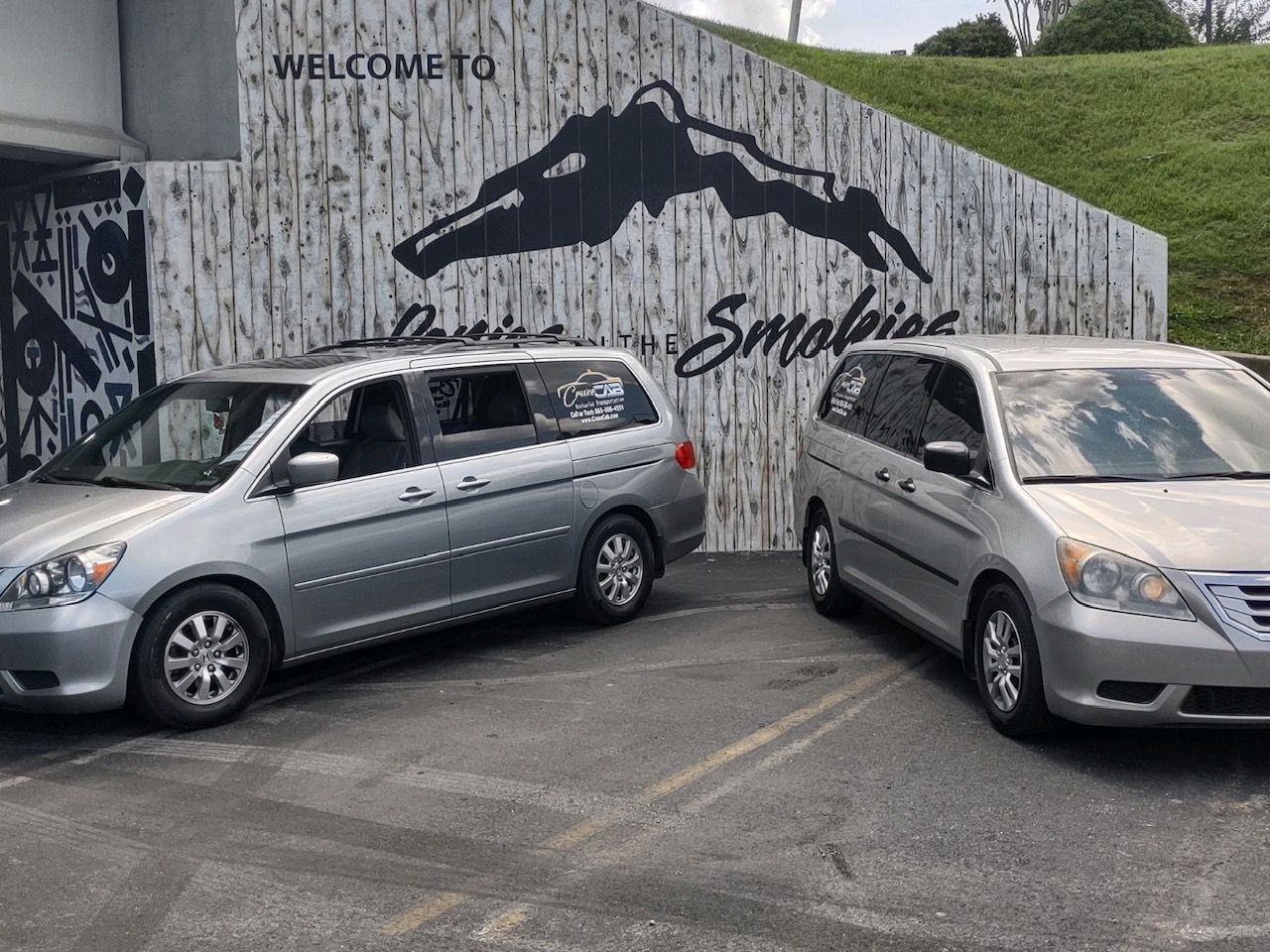two silver minivans are parked in a parking lot in front of a welcome sign .