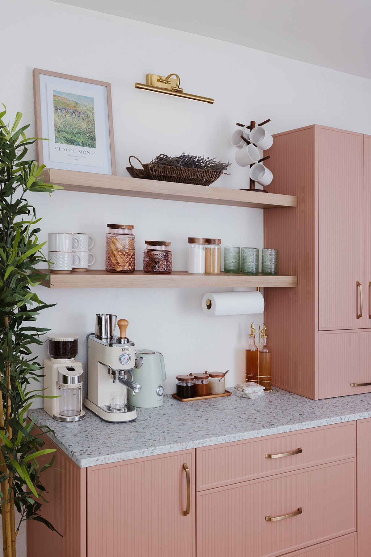 Pink kitchen counter with shelves, jars, coffee machine, and decorative items against a white wall.