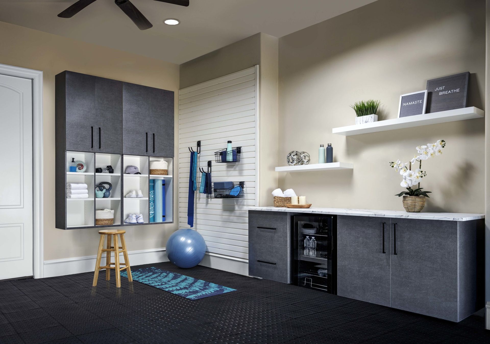 A home gym featuring dark grey cabinets, white wall shelves, a slat wall for equipment, a yoga mat, and an exercise ball.