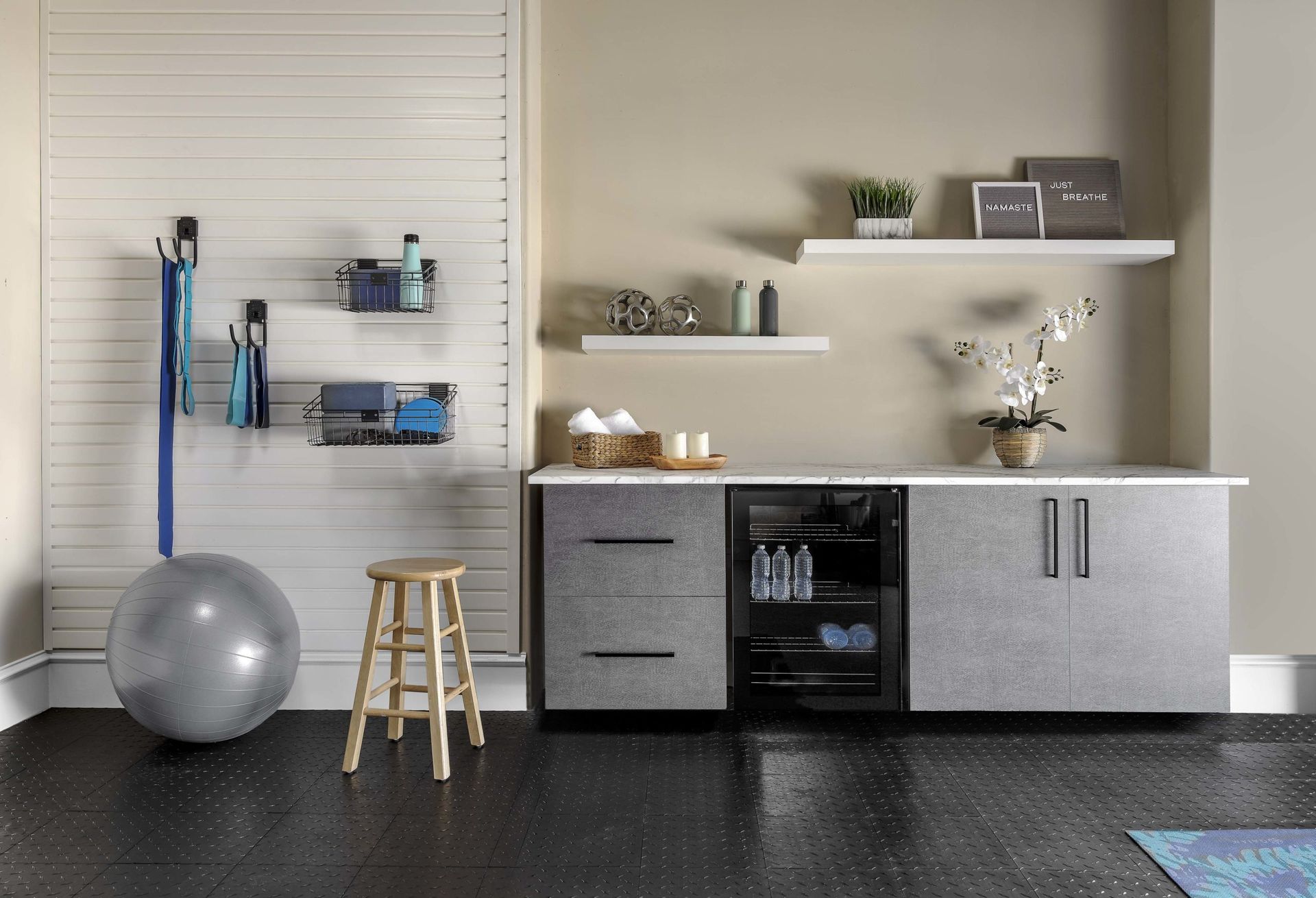 A home gym storage area with shelves, a gray cabinet with a mini-fridge, exercise ball, and wooden stool on black flooring.