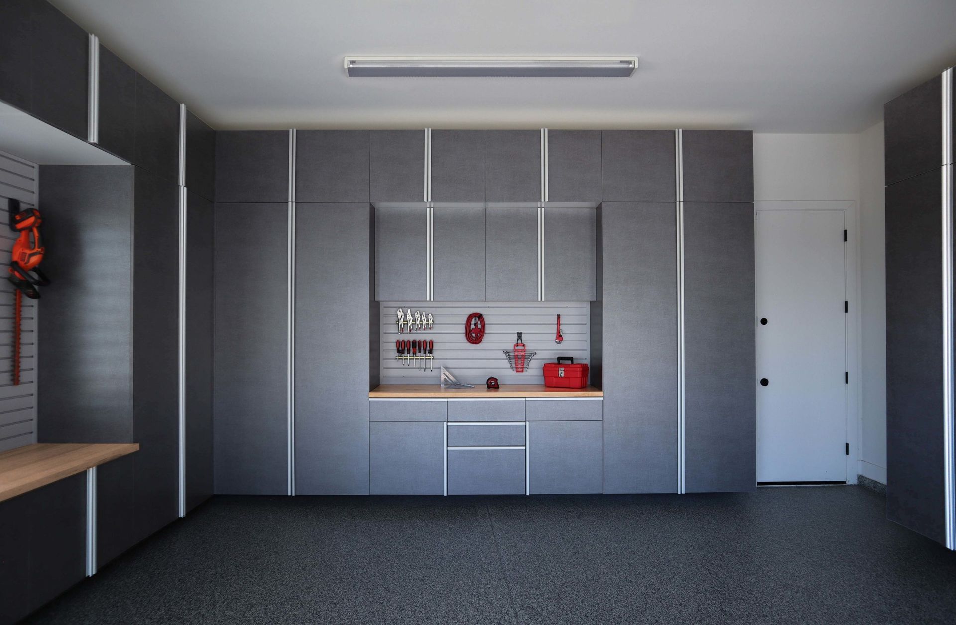 A modern garage featuring gray wall-to-wall cabinetry, a central workbench with tool storage, and a speckled floor.