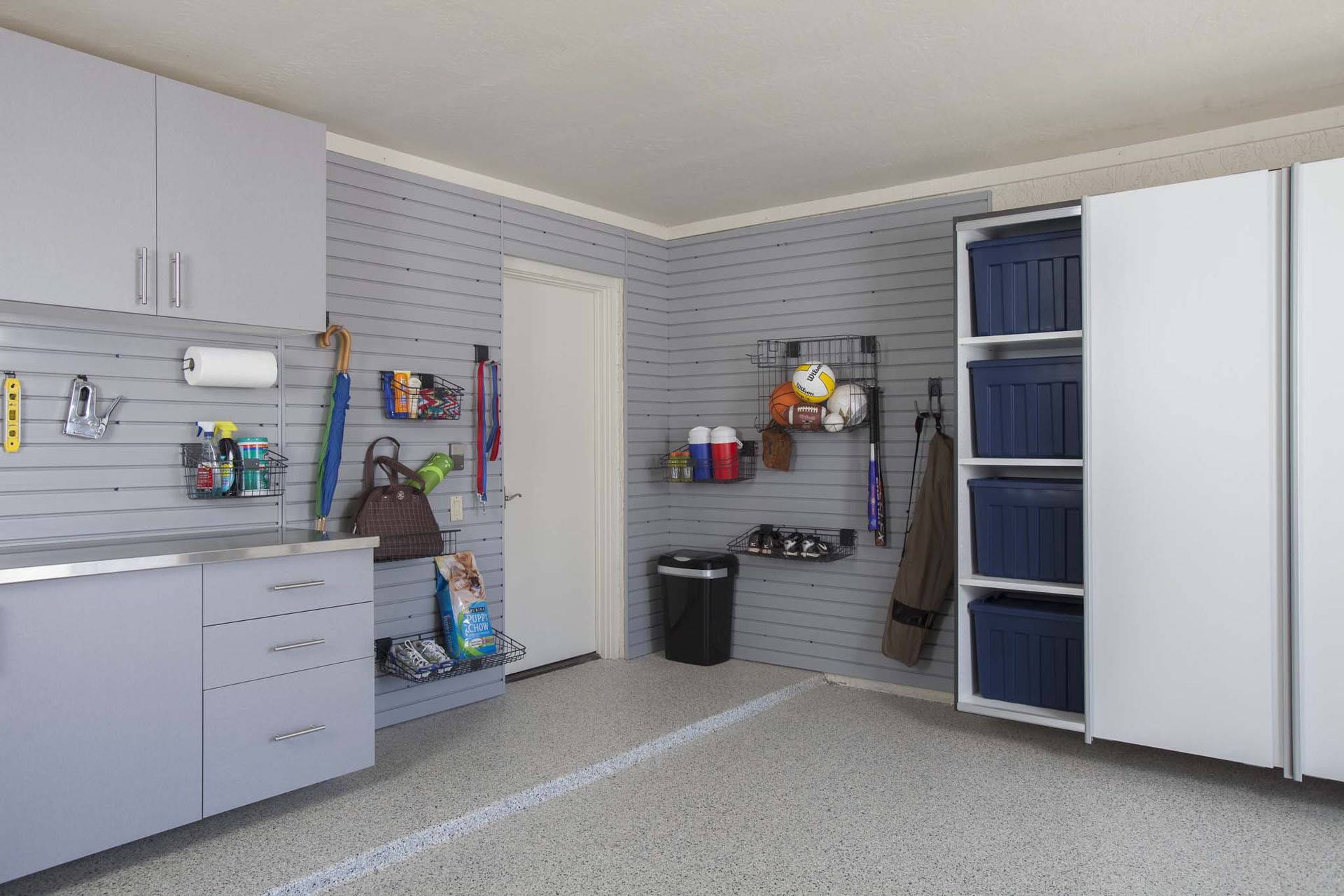 A well-organized garage with light gray slat walls, storage cabinets, shelving, and a tall cabinet with blue bins.