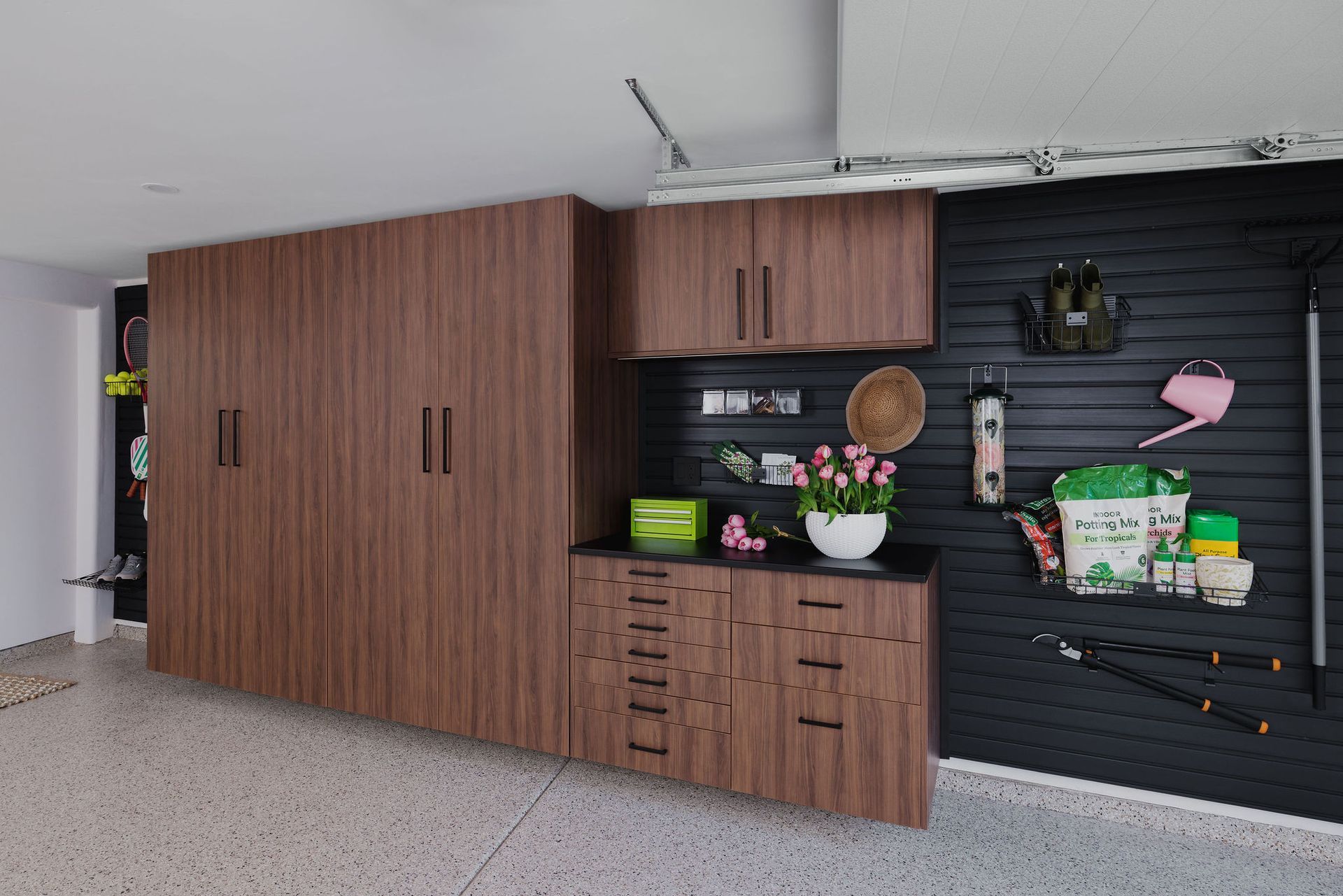 Modern wood-finish garage cabinetry with storage lockers, drawers, upper cabinets, and black slat-wall with tools.