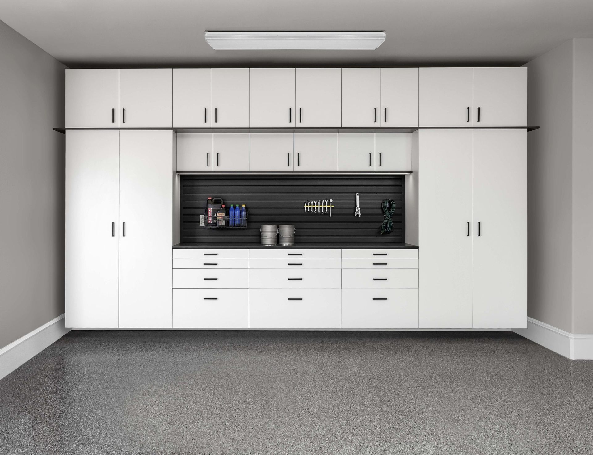 A wall-mounted white storage system with cabinets, drawers, and a black slatwall workspace in a garage.