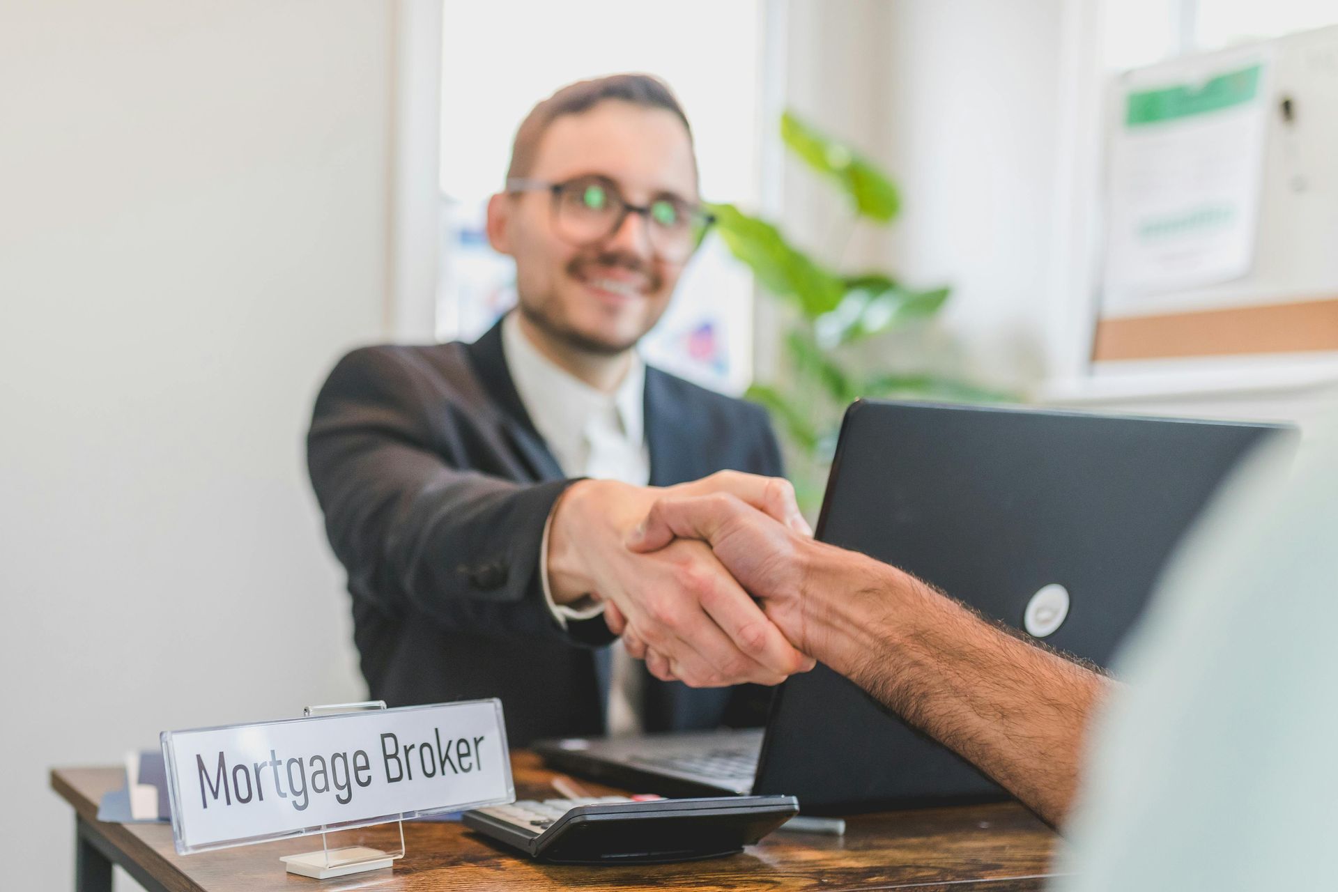 Mortgage broker shaking hands with a client at a desk; calculator, laptop, and sign that says “Mortgage Broker”.