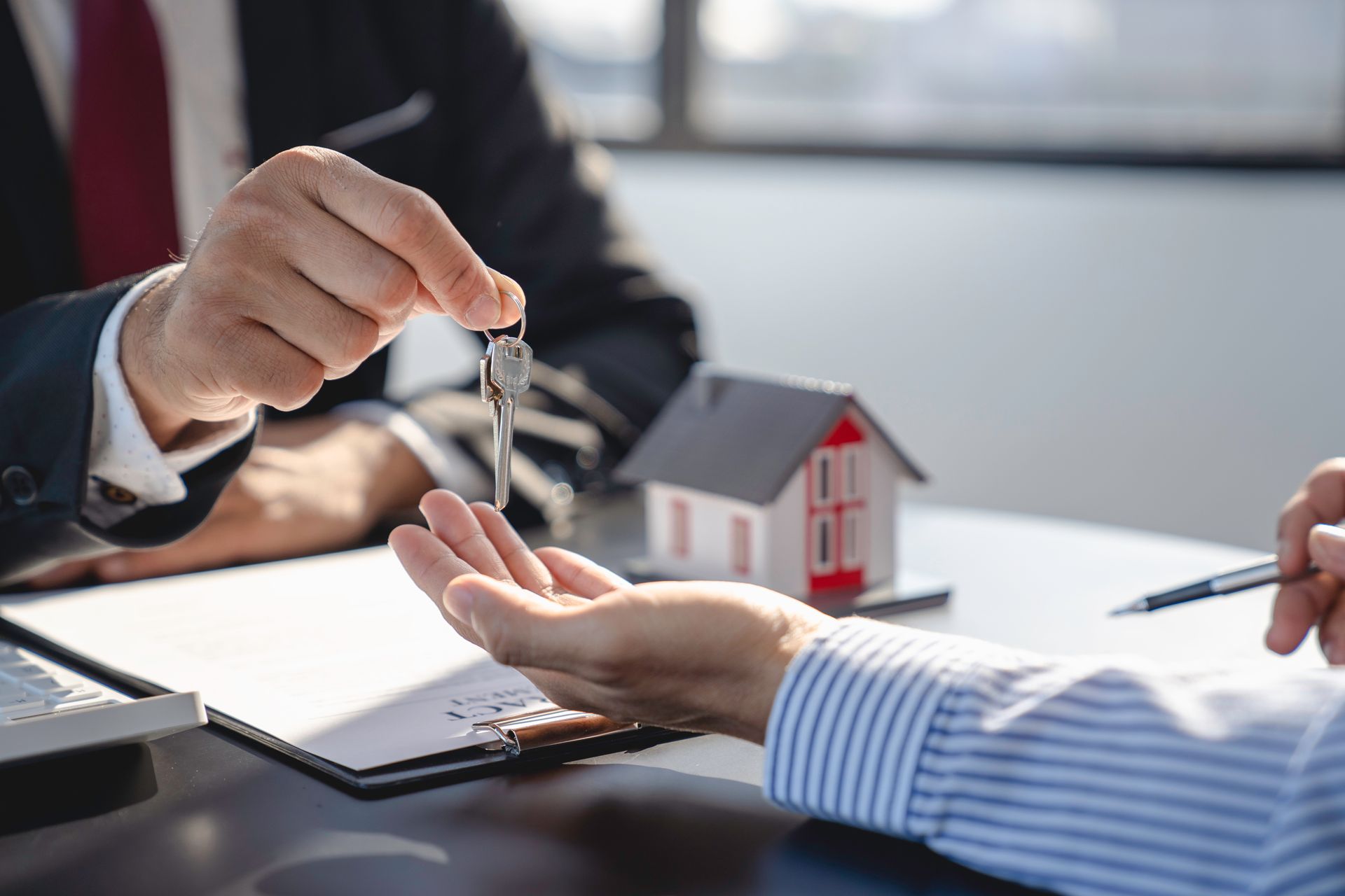 Person in suit hands keys to another person; miniature house model sits on table.