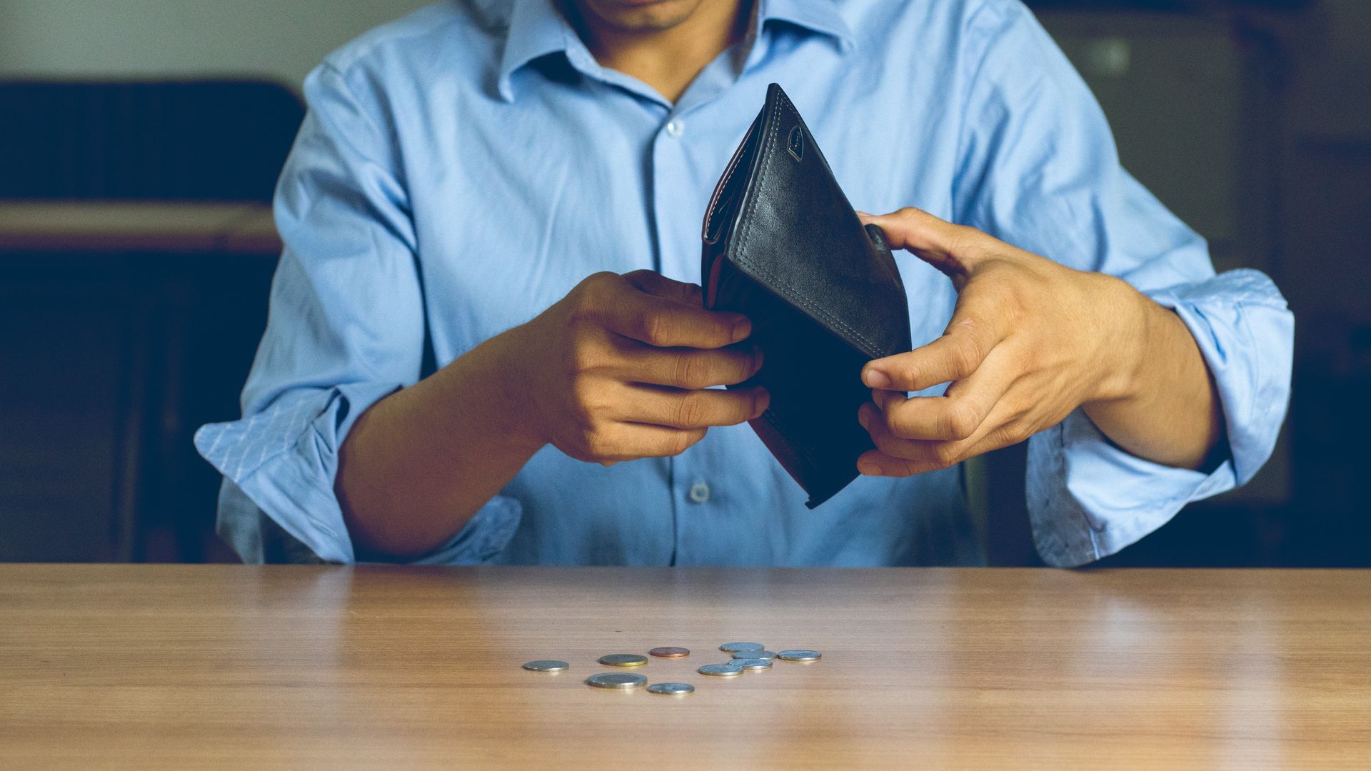 Man in blue shirt, opening an empty wallet, coins on table.