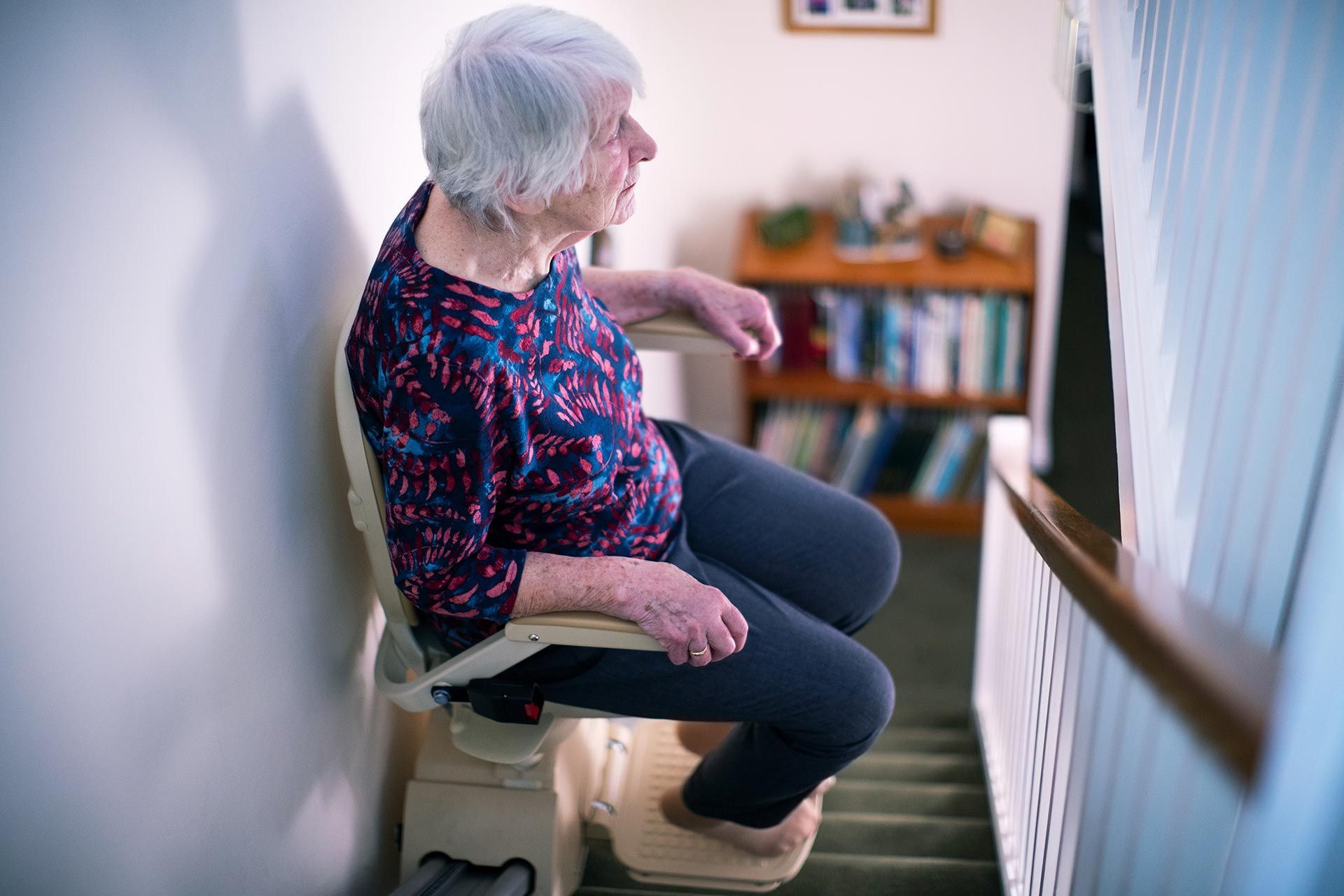 Person using a stairlift on a home staircase beside railing and bookshelf.