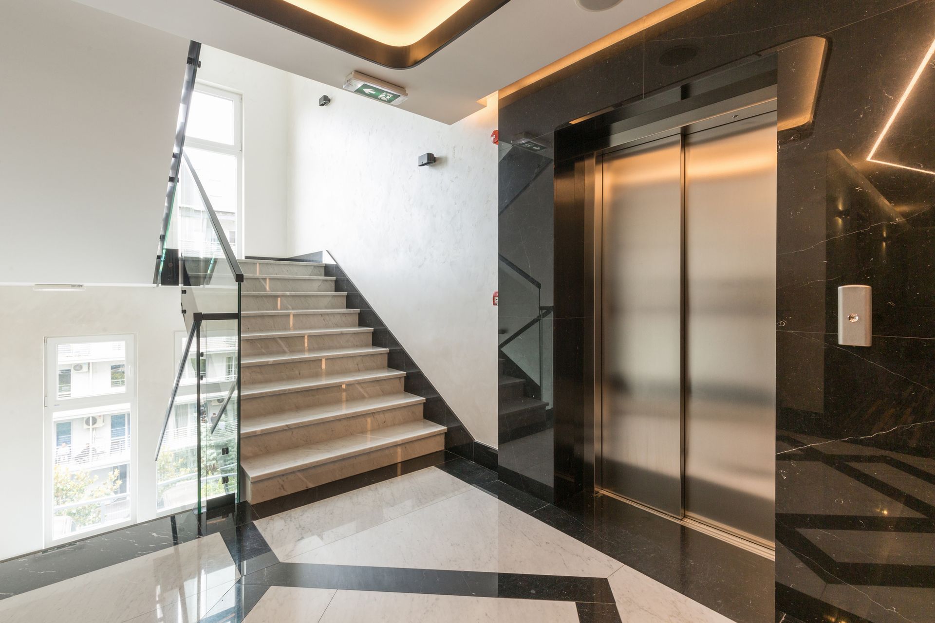 Staircase with glass railing, elevator, and white walls in a well-lit modern building.