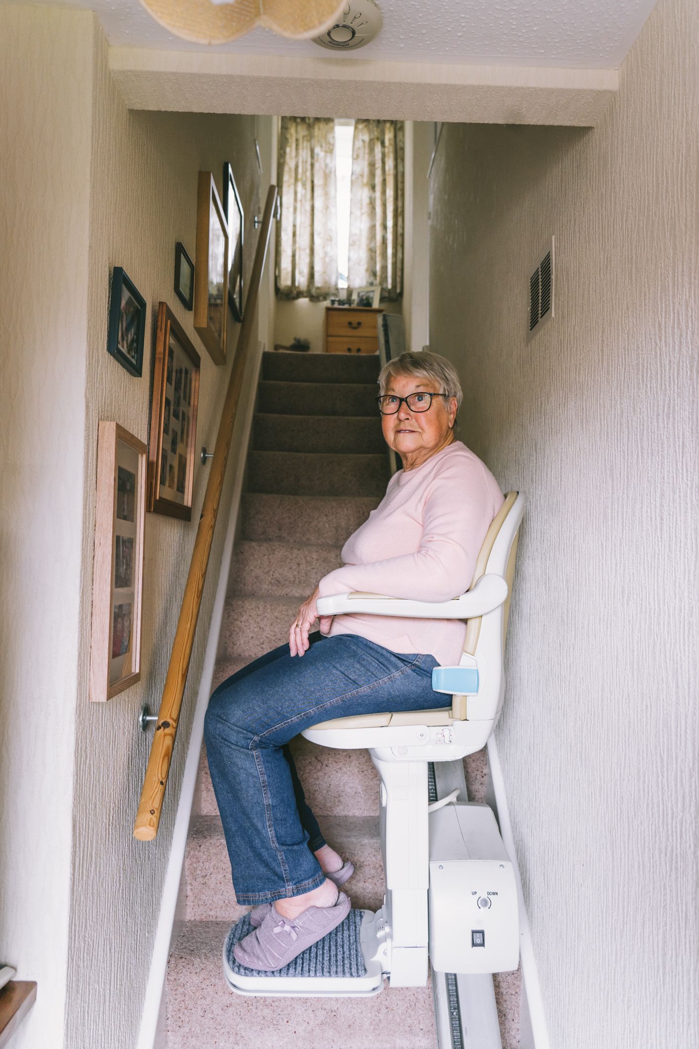 Senior woman with pink sweater sits in a home stair lift in front of a staircase inside a home.