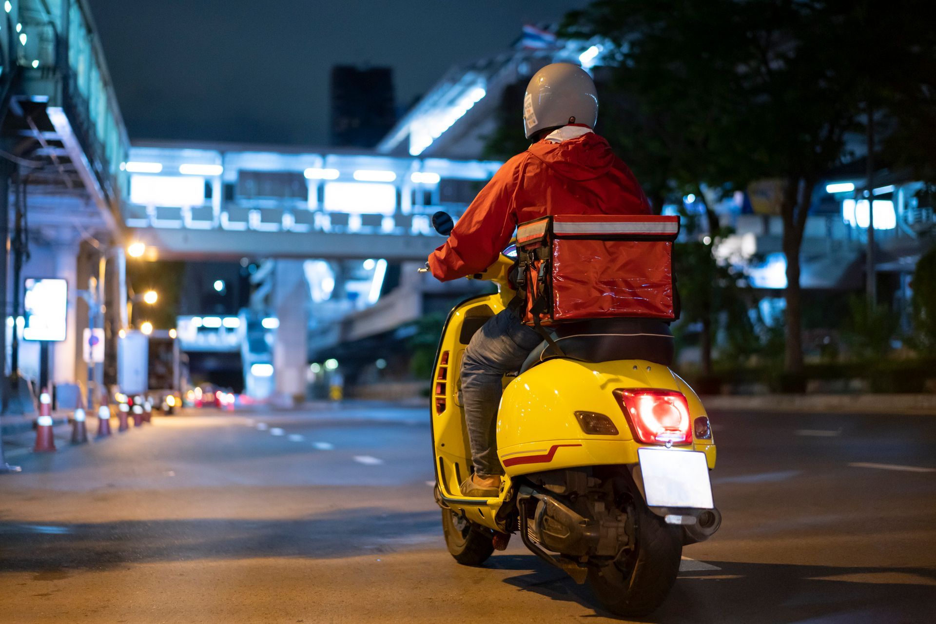 A person on a yellow scooter delivering food at night, wearing a red jacket and helmet. A person on a yellow scooter delivering food at night, wearing a red jacket and helmet.