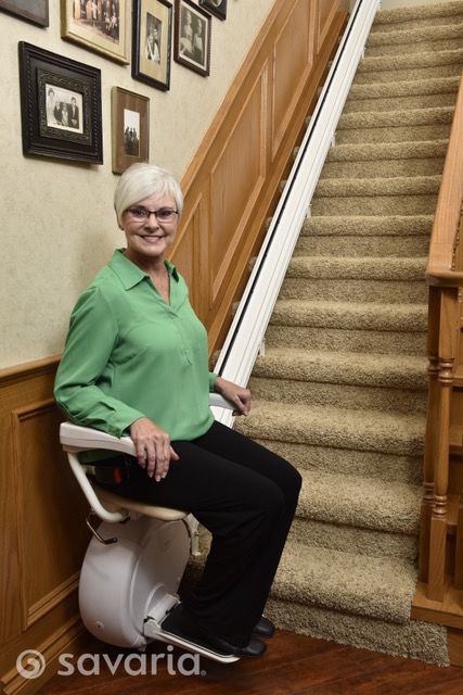 Elderly woman sitting on a stair lift at the bottom of a carpeted staircase in a home.