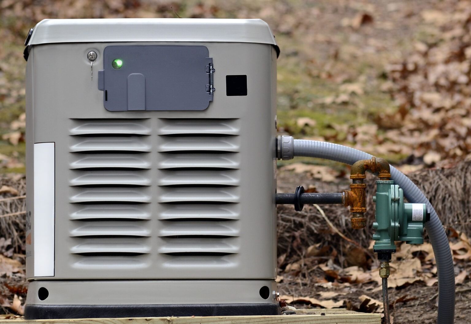 Gray outdoor generator with vented front and connected hoses on a gravel surface among leaves