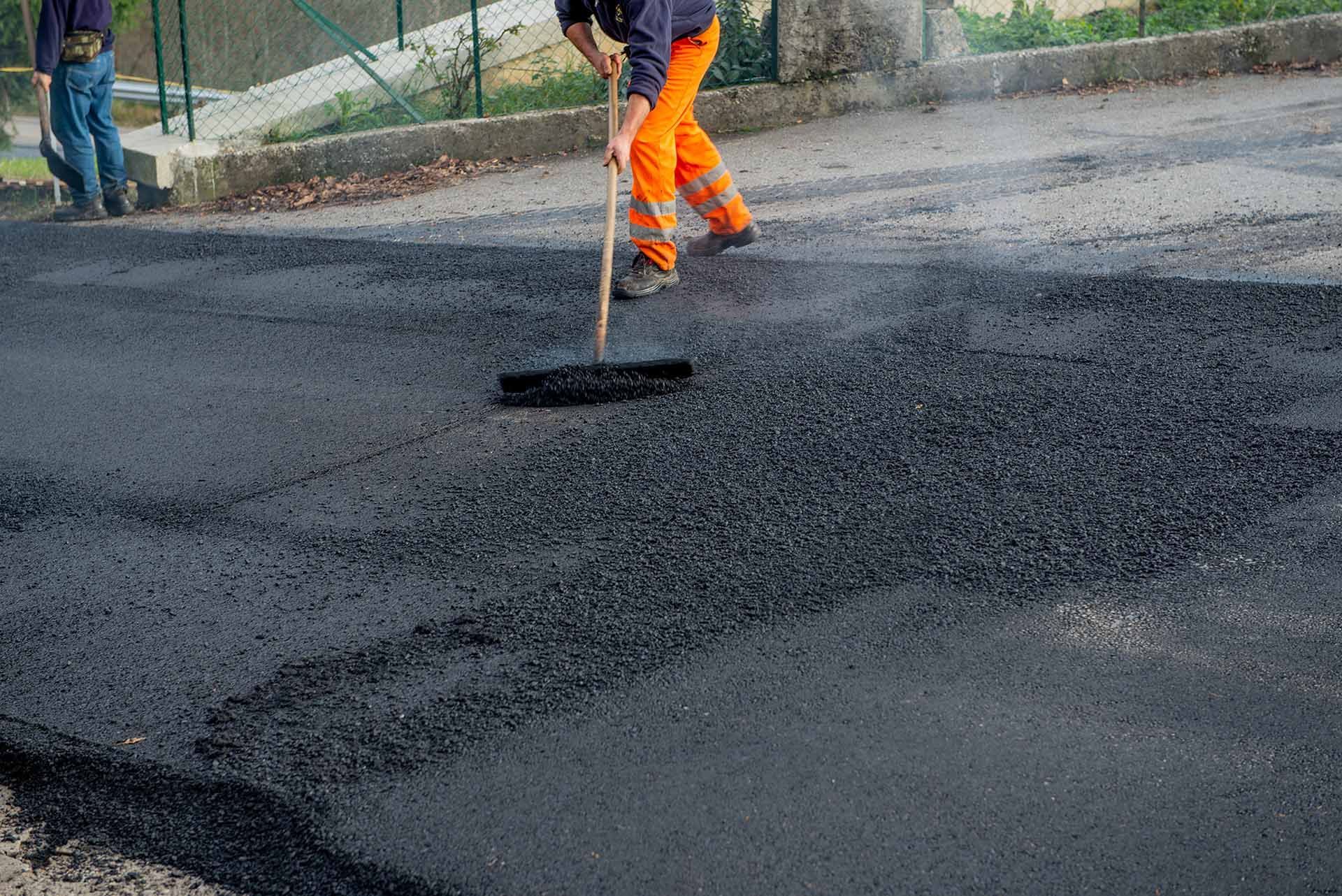 Worker spreading fresh asphalt with a rake during road resurfacing on a paved street.
