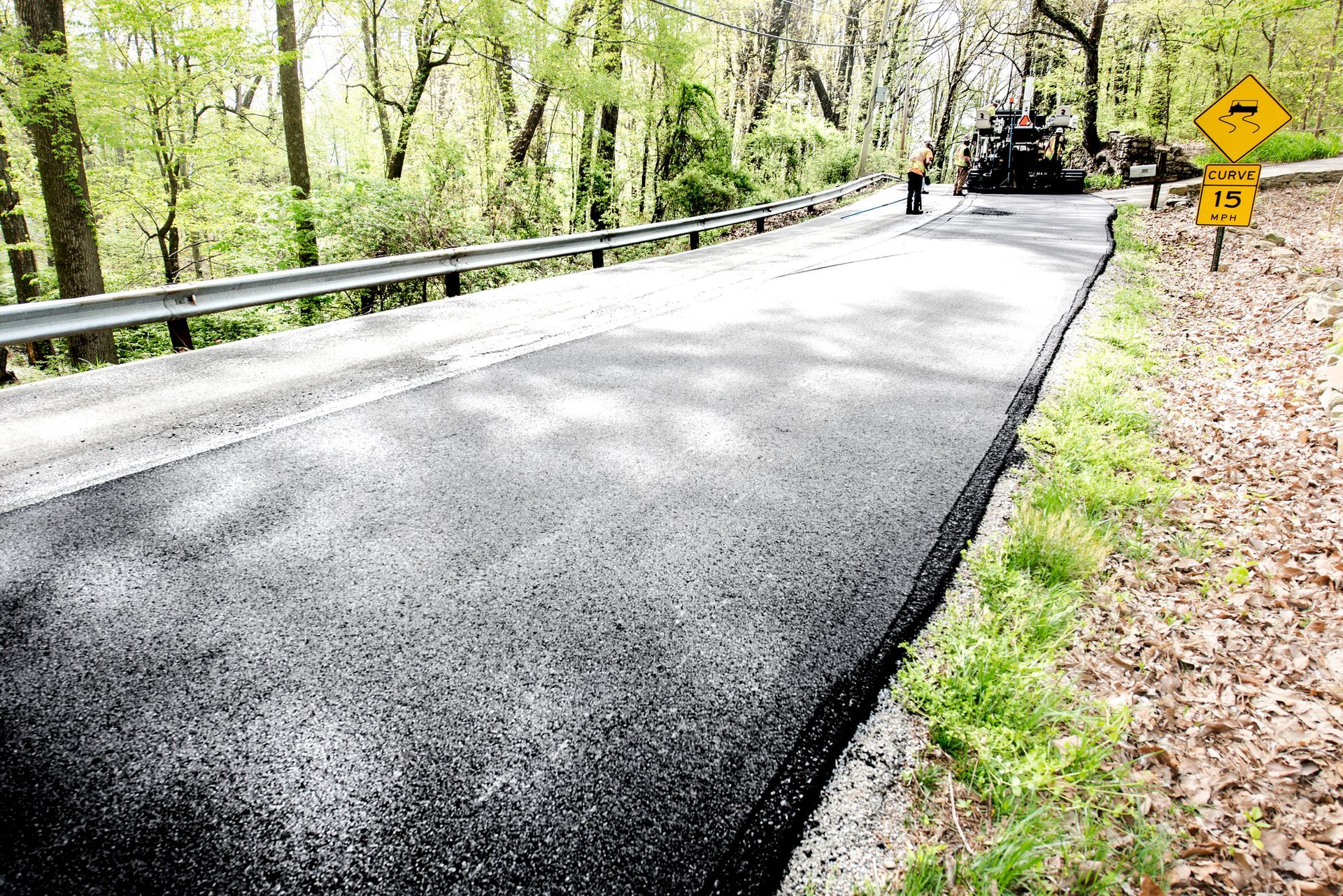 Fresh asphalt road in wooded area with paving gear and a curve warning sign nearby.