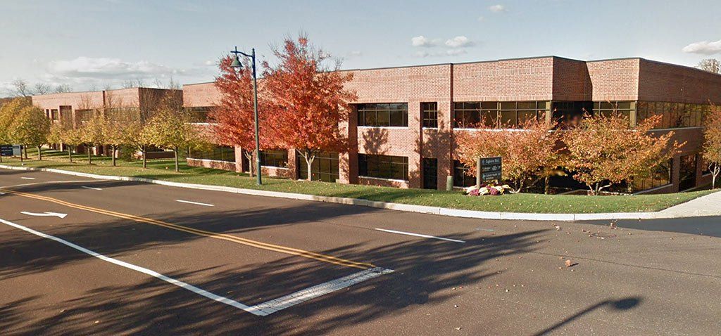 A two-story brick building with trees in autumn; a parking lot and street in front.