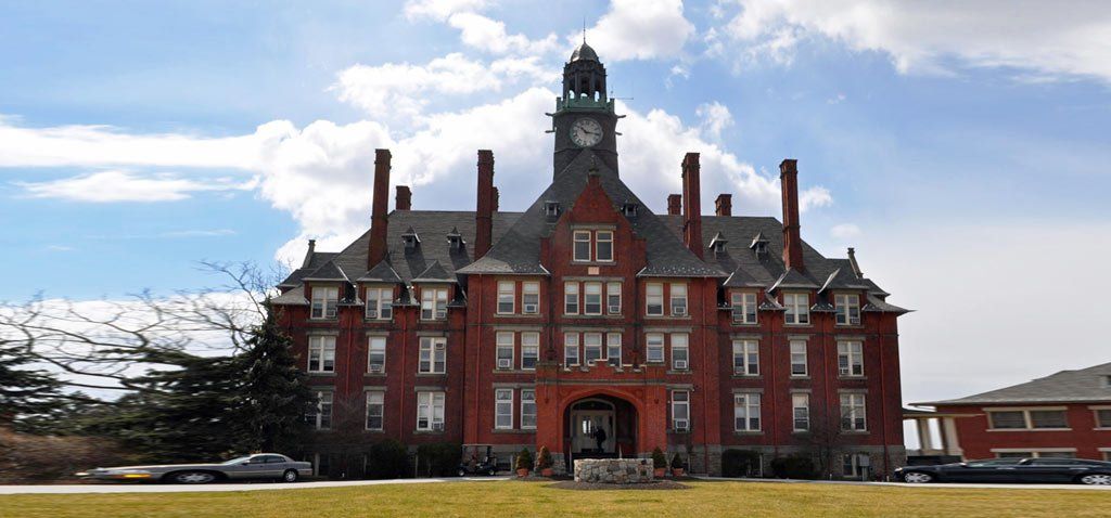 Red brick building with clock tower under a cloudy sky, set on a grassy field.
