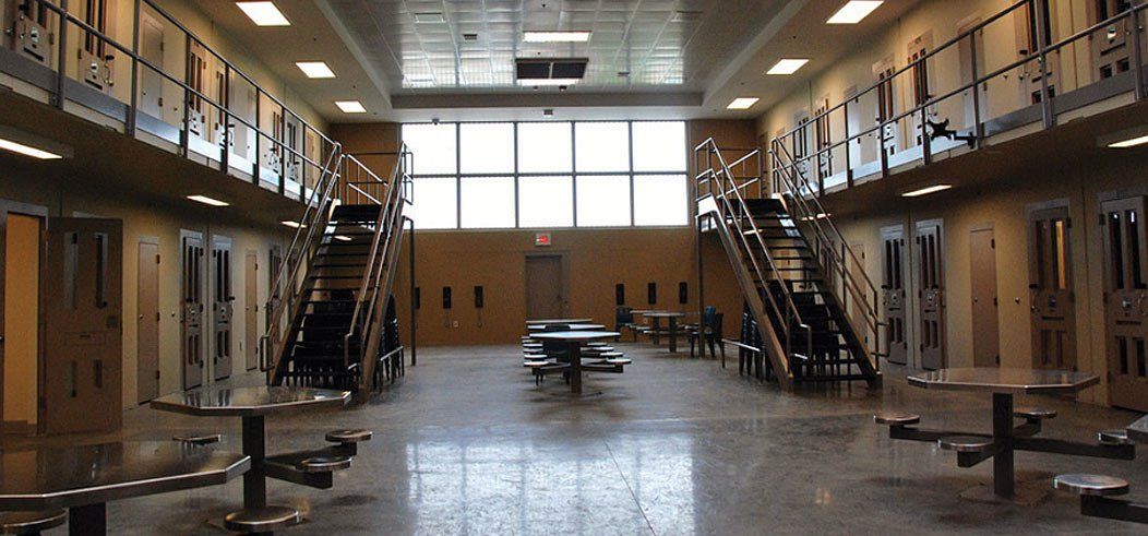 Interior of a prison with cells lining the walls, stairs, and tables in a central area.