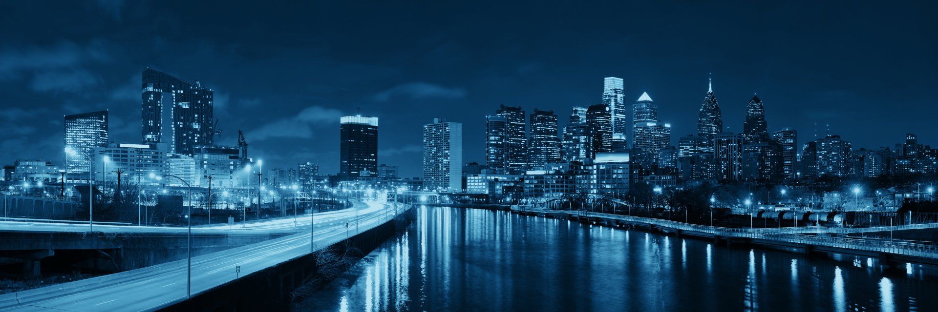 Nighttime view of a city skyline with building lights reflecting in a river, all in blue tones.