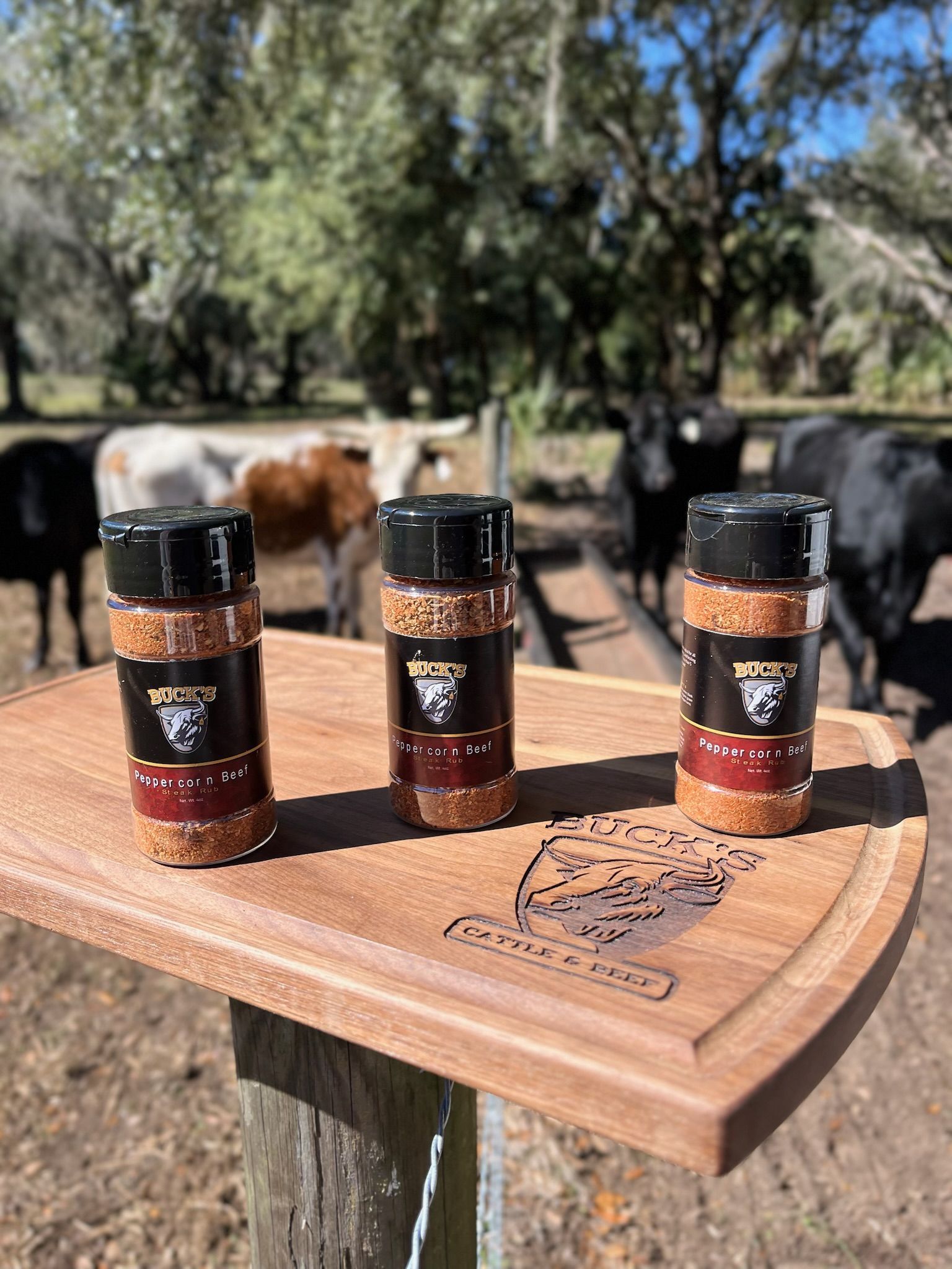 Three spice jars on a wooden table outdoors, with trees and livestock blurred in the background.
