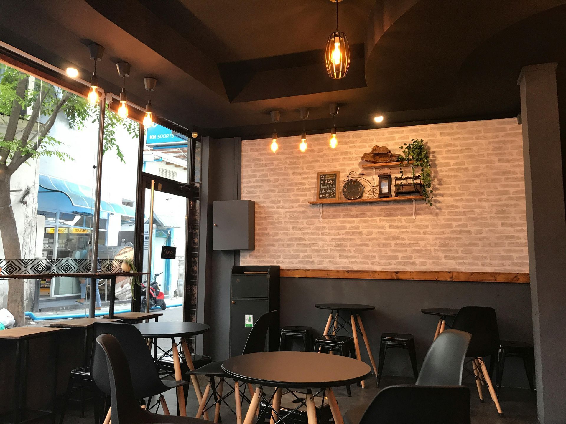 Interior of a cafe: tables and chairs, brick accent wall with decor, large window, hanging lights.