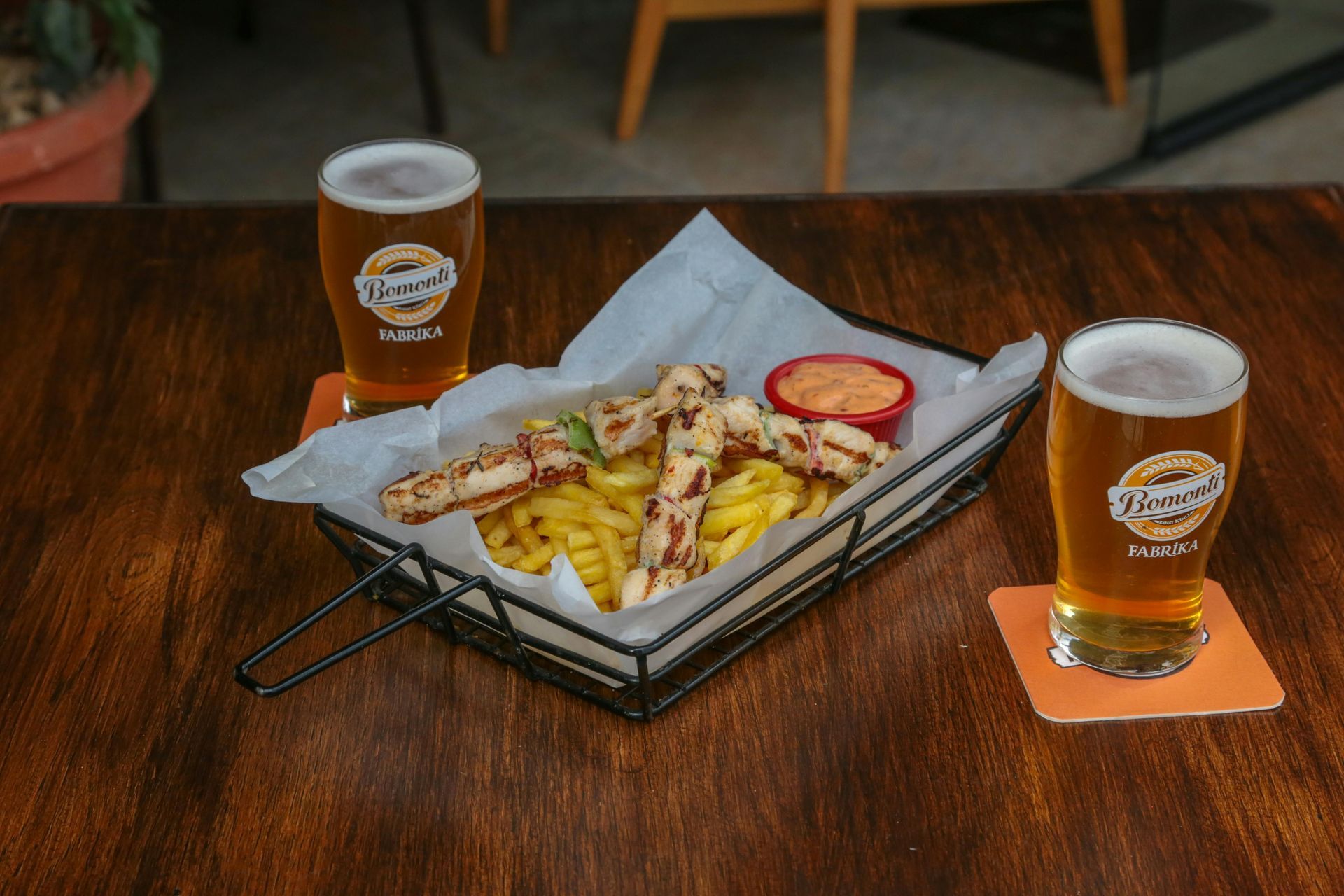 Two beers, fries, and grilled food on a basket, wooden table.