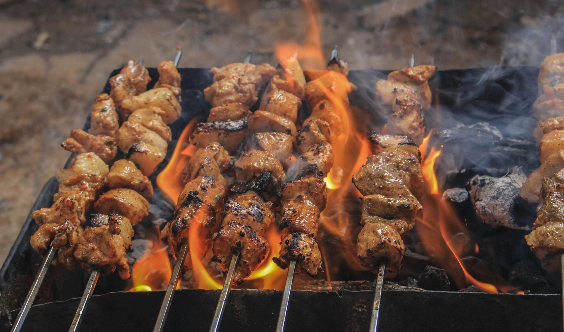 Skewers of meat on fire cooking on a charcoal grill.
