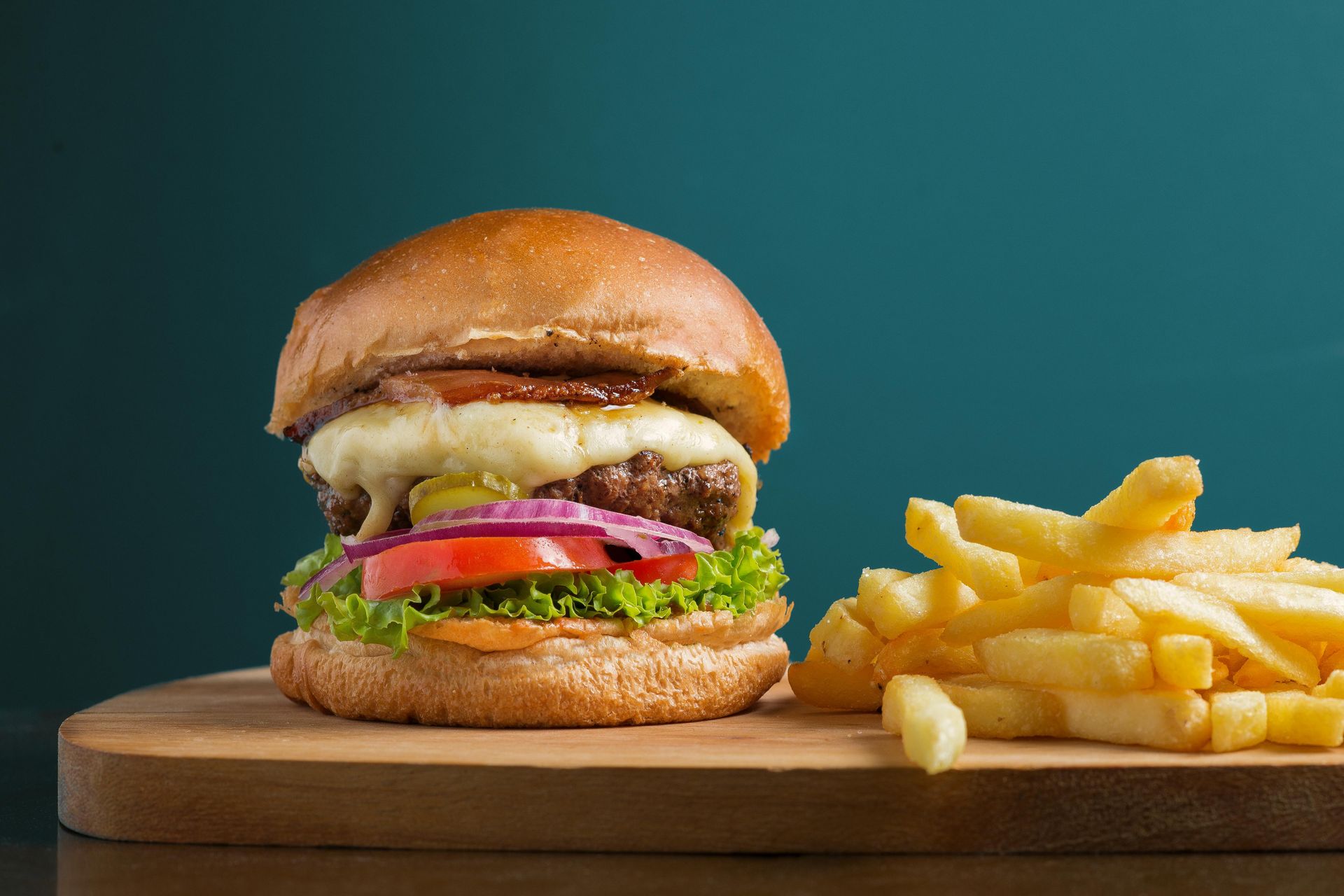 Burger with fries on a wooden board; green lettuce, red onion, tomato, cheese, bun.