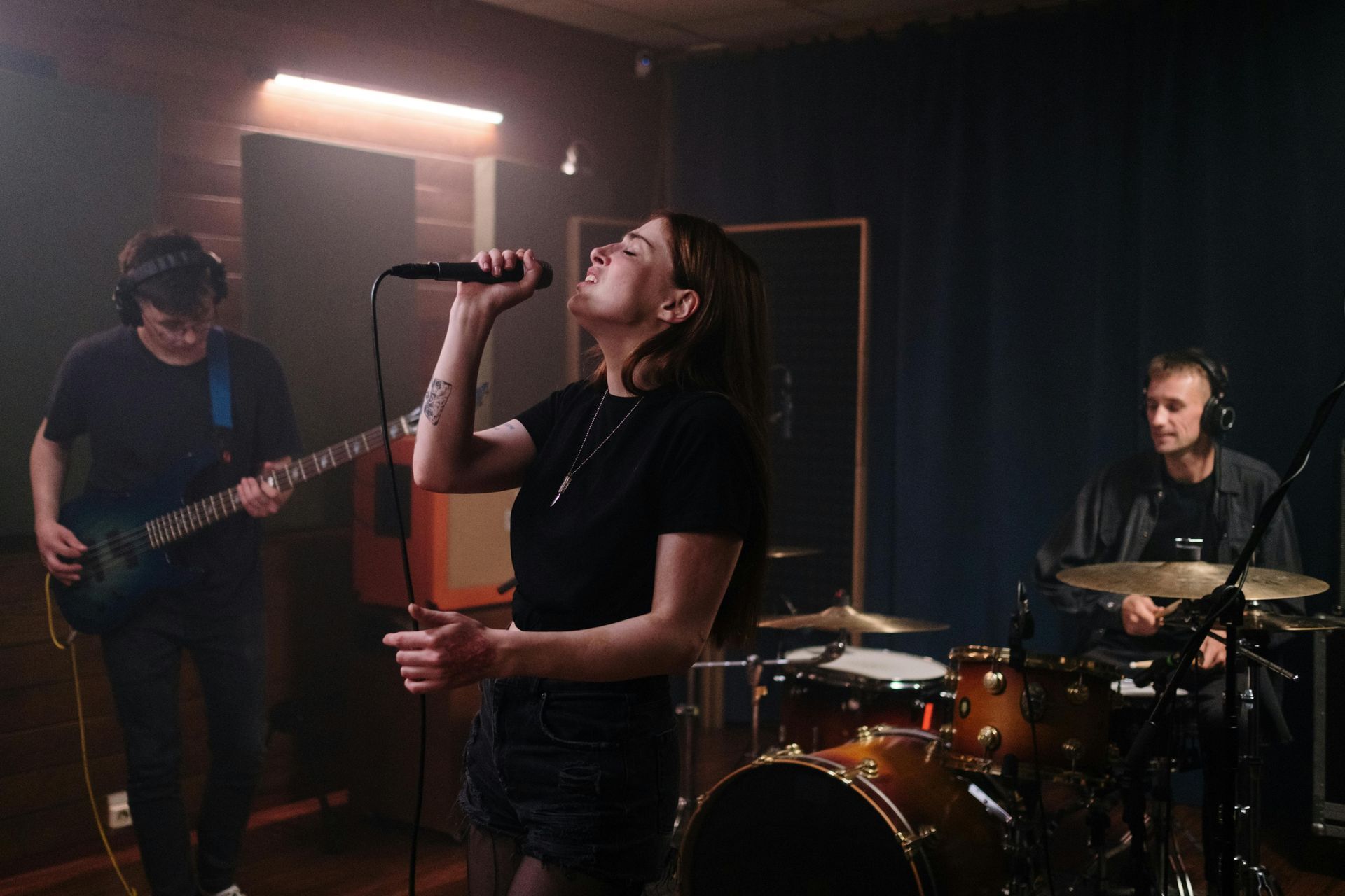 A band performs in a studio: a female singer, guitarist, and drummer. Dim lighting and smoky atmosphere.