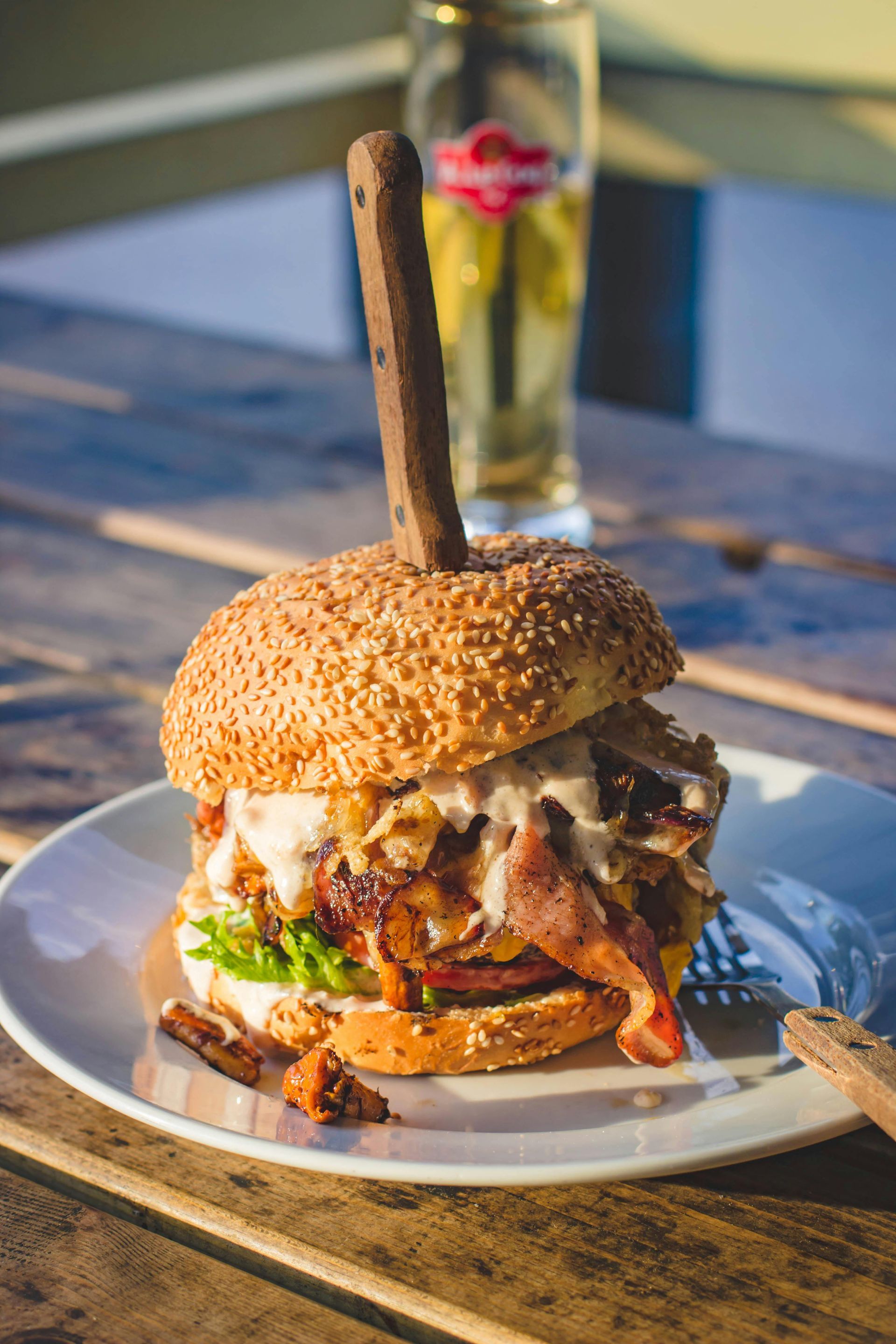 Sesame seed burger impaled with a knife, with bacon and toppings, on a plate next to a glass of beer.