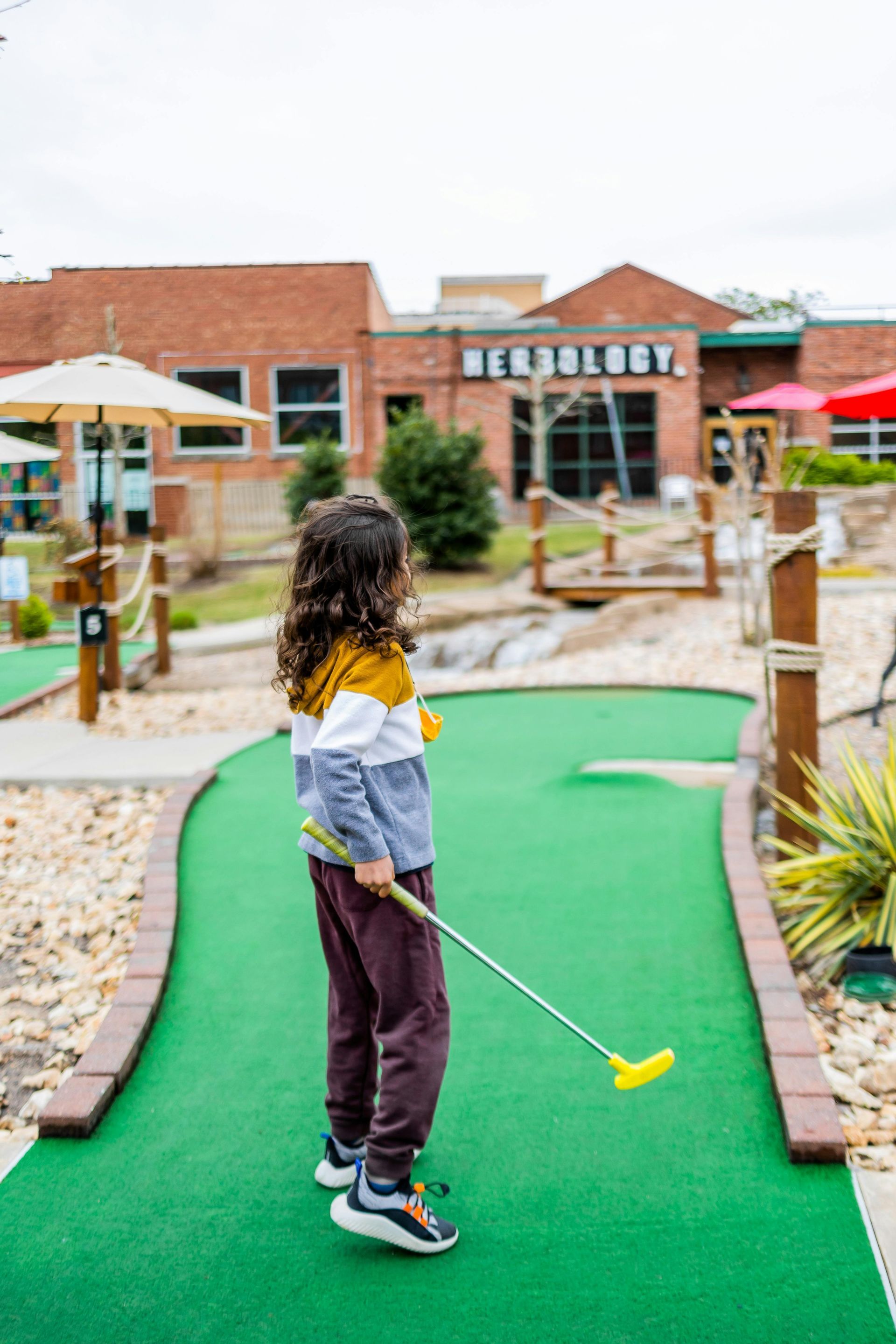Young child with curly hair plays miniature golf on a green course; brick building in background.