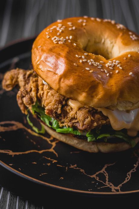 Fried chicken sandwich on a sesame bagel with lettuce and sauce, served on a black plate.
