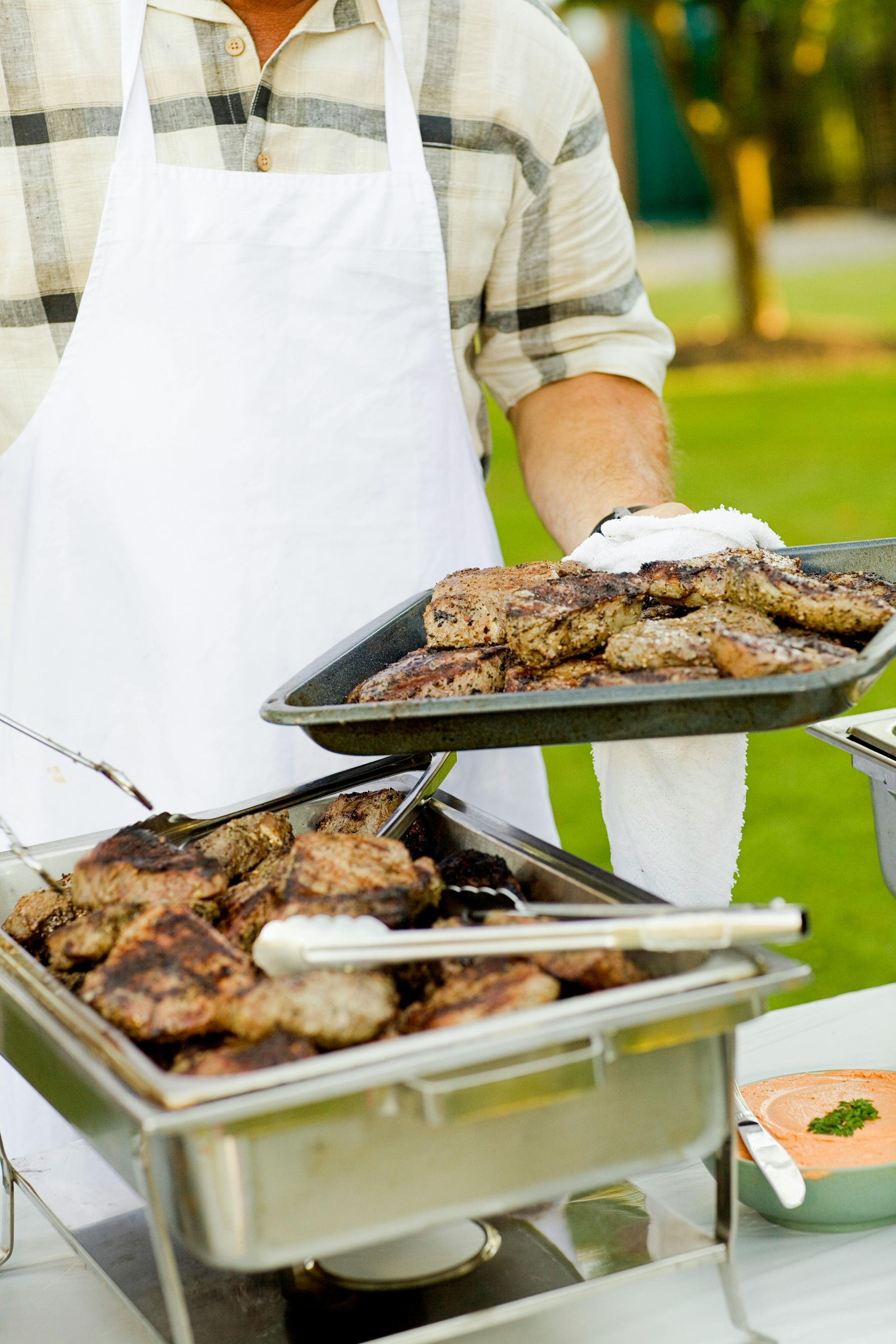 Person in white apron holding a tray of grilled meat, serving food at an outdoor gathering.