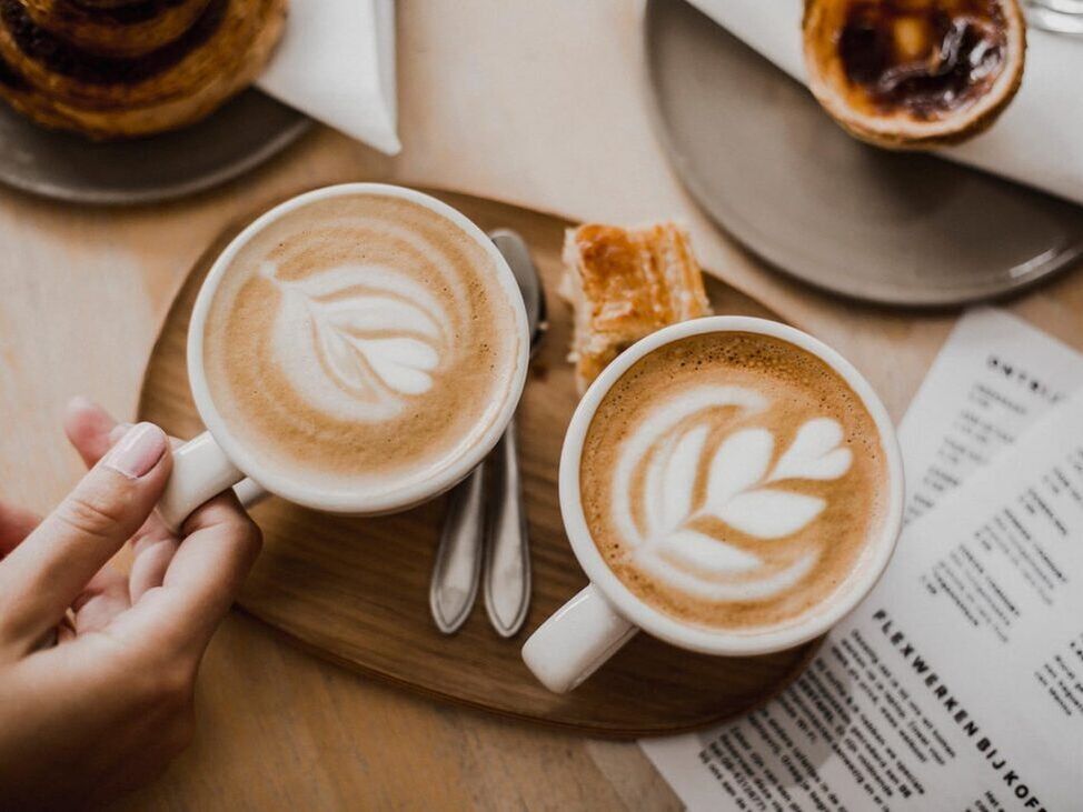 Two lattes with leaf art on a wooden tray, hand holding one. Pastries and menu are nearby.