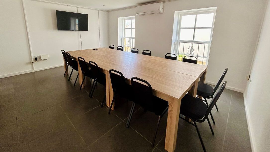 A long wooden conference table with black chairs in a bright room with windows and a wall-mounted TV.