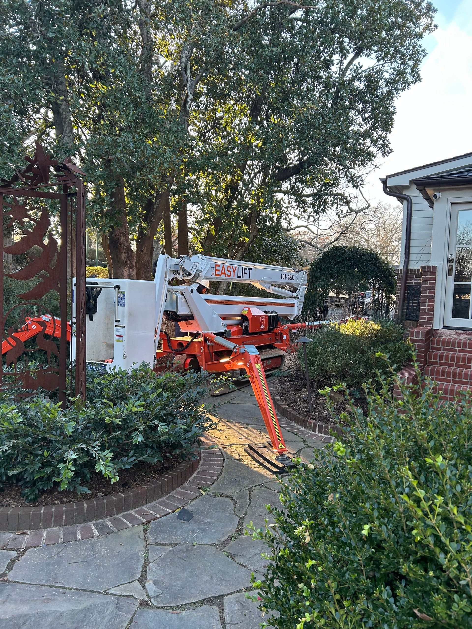 Cherry picker in yard near a brick house, likely trimming trees. Orange and white machinery.
