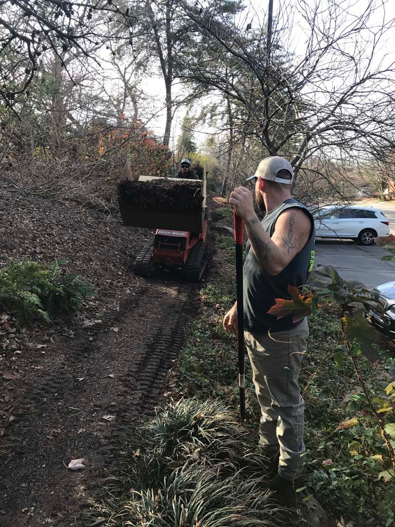 Man with shovel on a wooded trail, another person operates a small vehicle in the background.