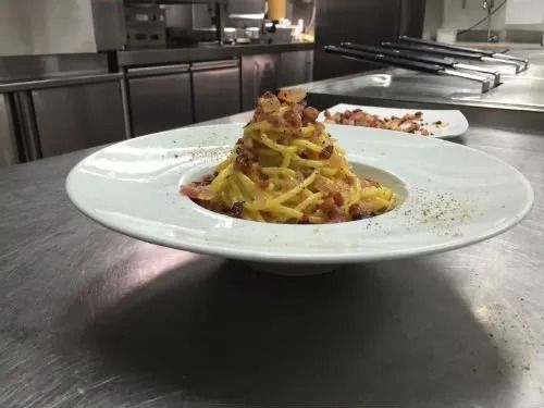 Un piatto bianco con sopra noodles e pancetta su un bancone.