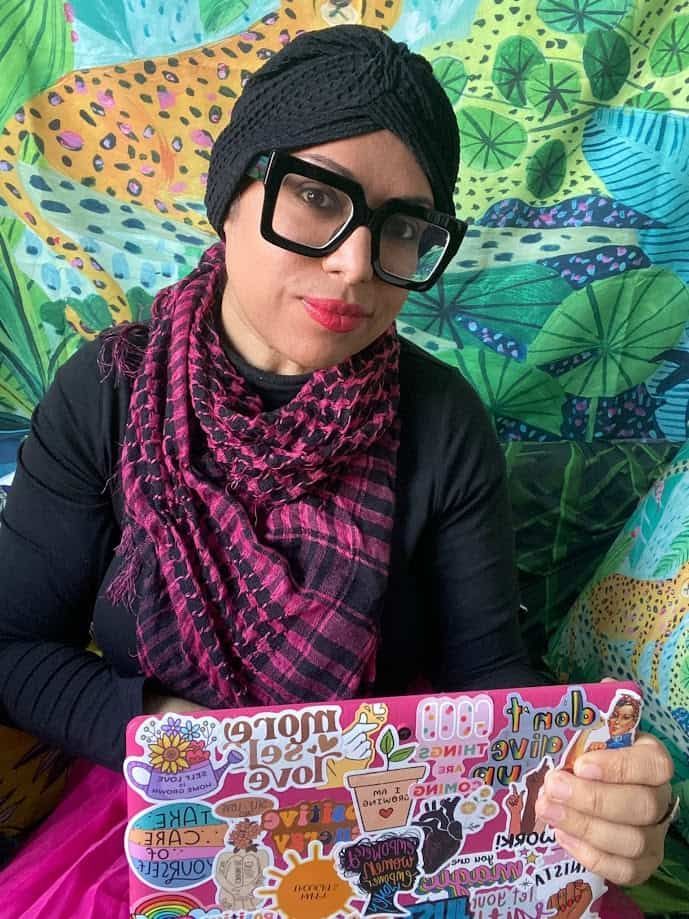Woman with glasses and headscarf holding a laptop with stickers; colorful background.