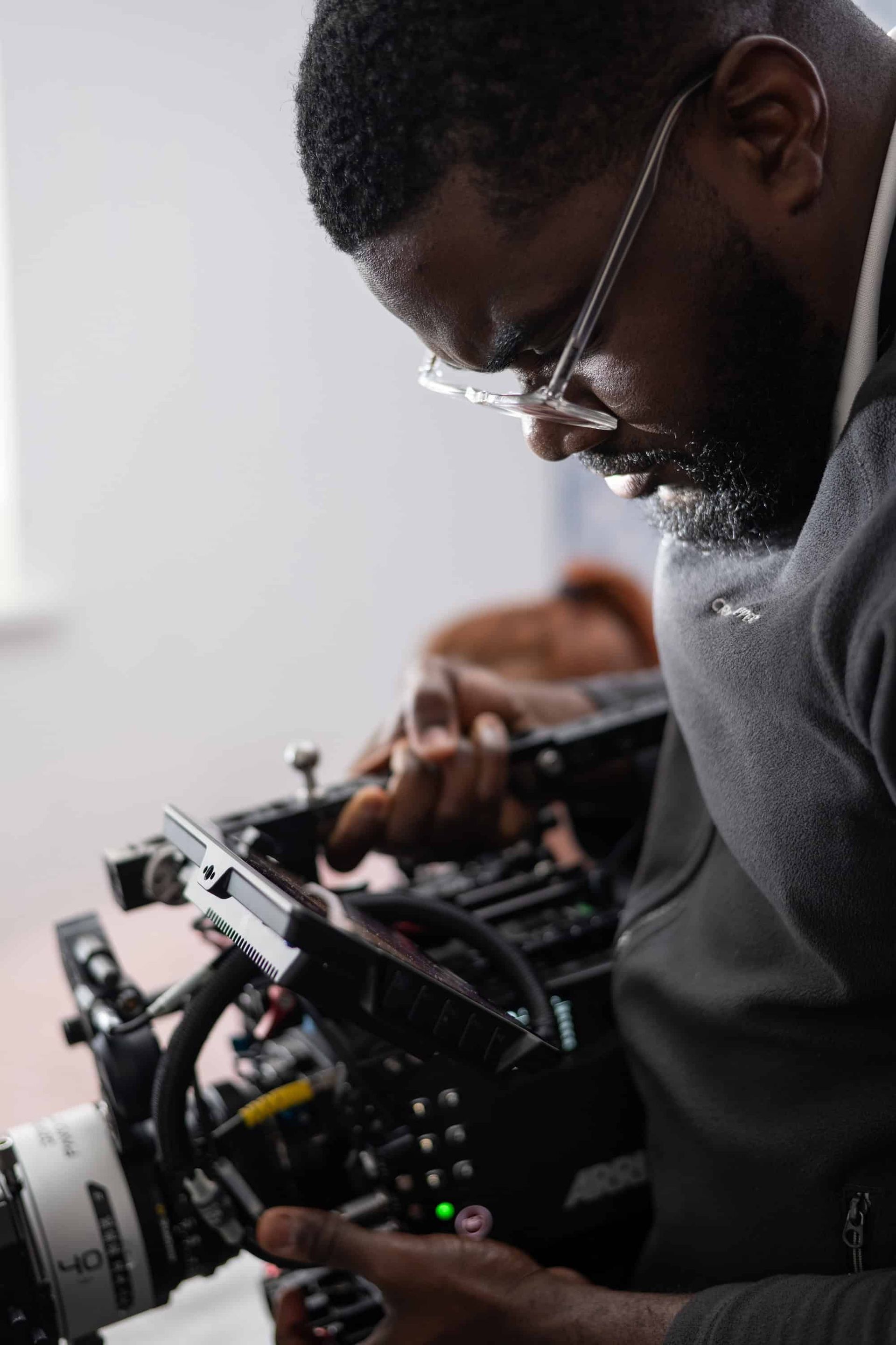 Black man wearing glasses, filming with a professional camera indoors.