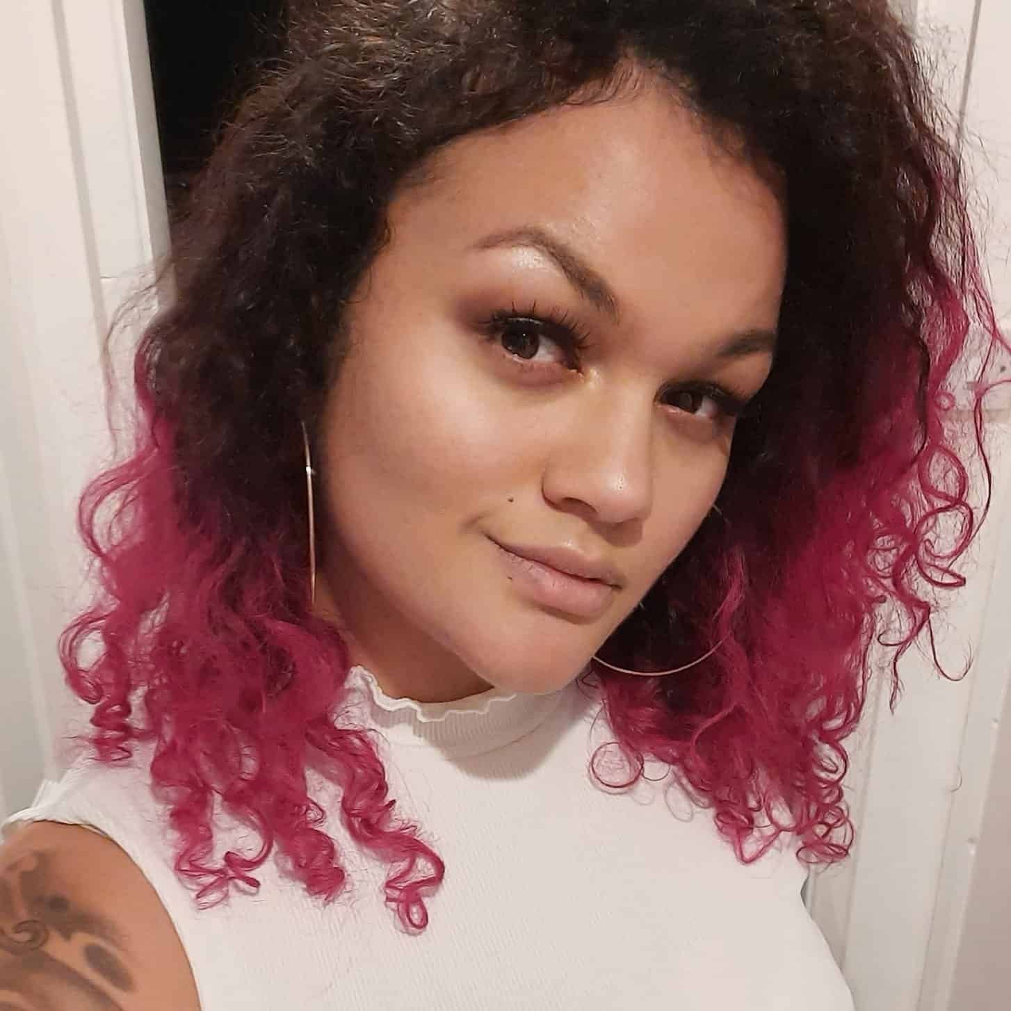 Woman with curly hair, pink highlights, white top, hoop earrings, smiling, looking at the camera.