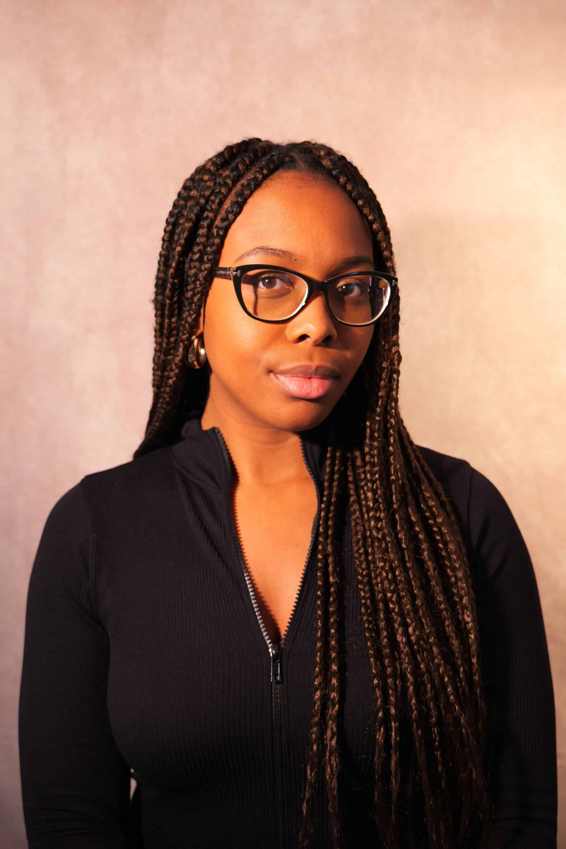 Young Black woman with long braids, glasses, and black zip-up. Pale background.