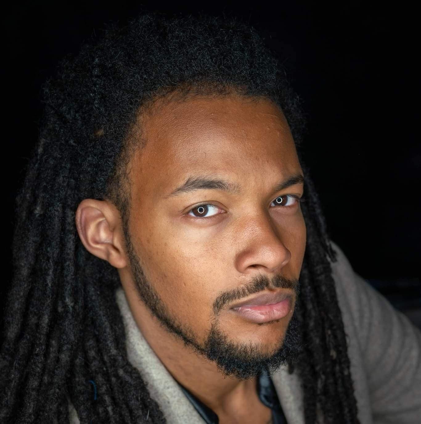 A Black man with dreadlocks and a goatee gazes toward the camera, with a neutral expression.