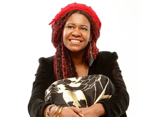 Woman with red hat smiles, holding a patterned pillow. Black jacket, gold bracelets, white background.