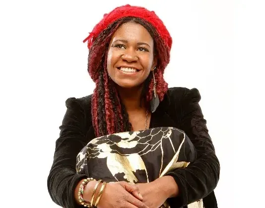 Woman with red and black braids and hat, smiling, holding patterned fabric, wearing bracelets.