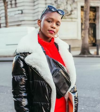 Woman with short hair wearing black jacket with white fur trim, red sweater, sunglasses, and gold hoop earrings, standing outside.